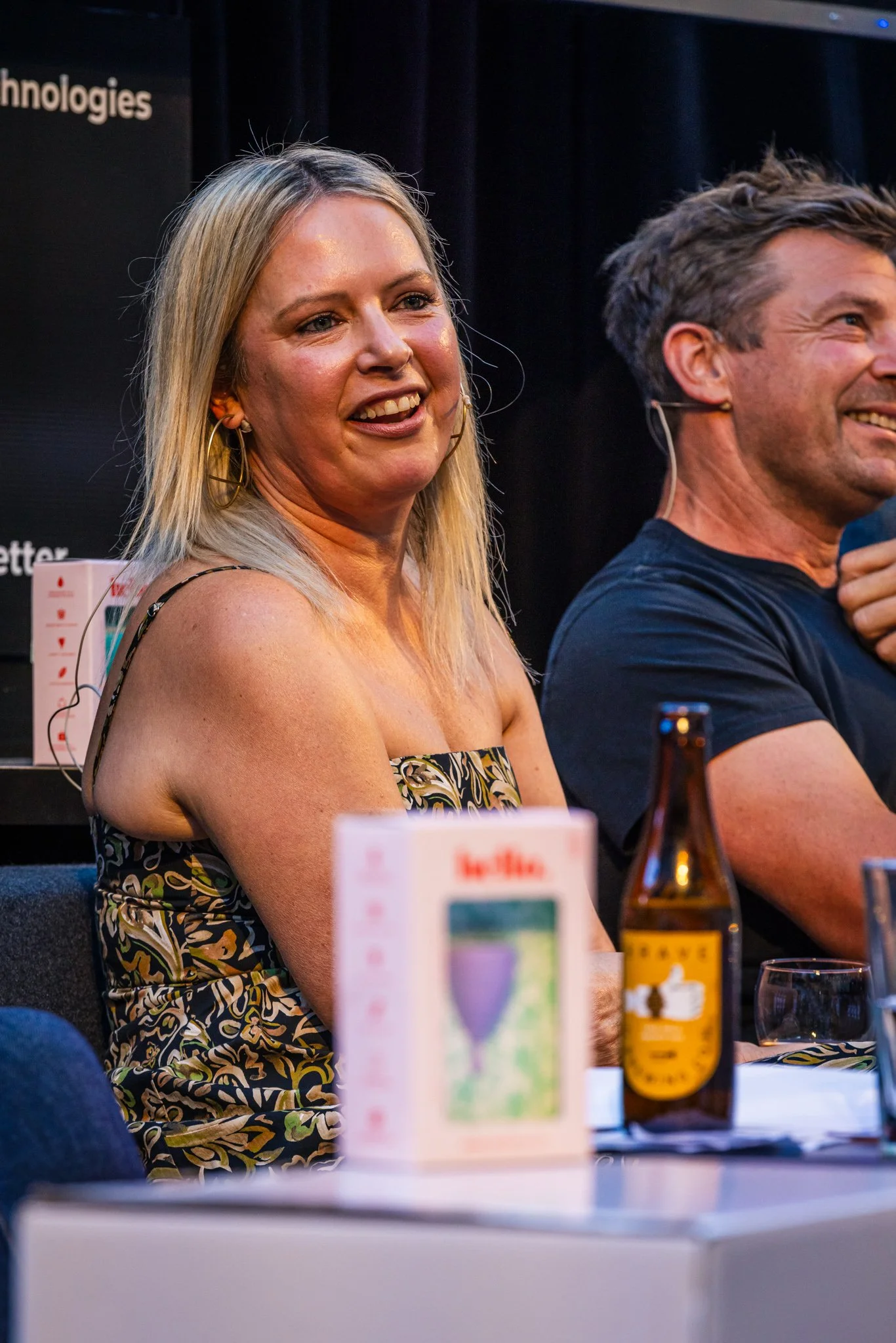 A woman with blonde hair smiling and wearing a patterned dress, seated at a table during a conference or panel discussion, with a man beside her also smiling.