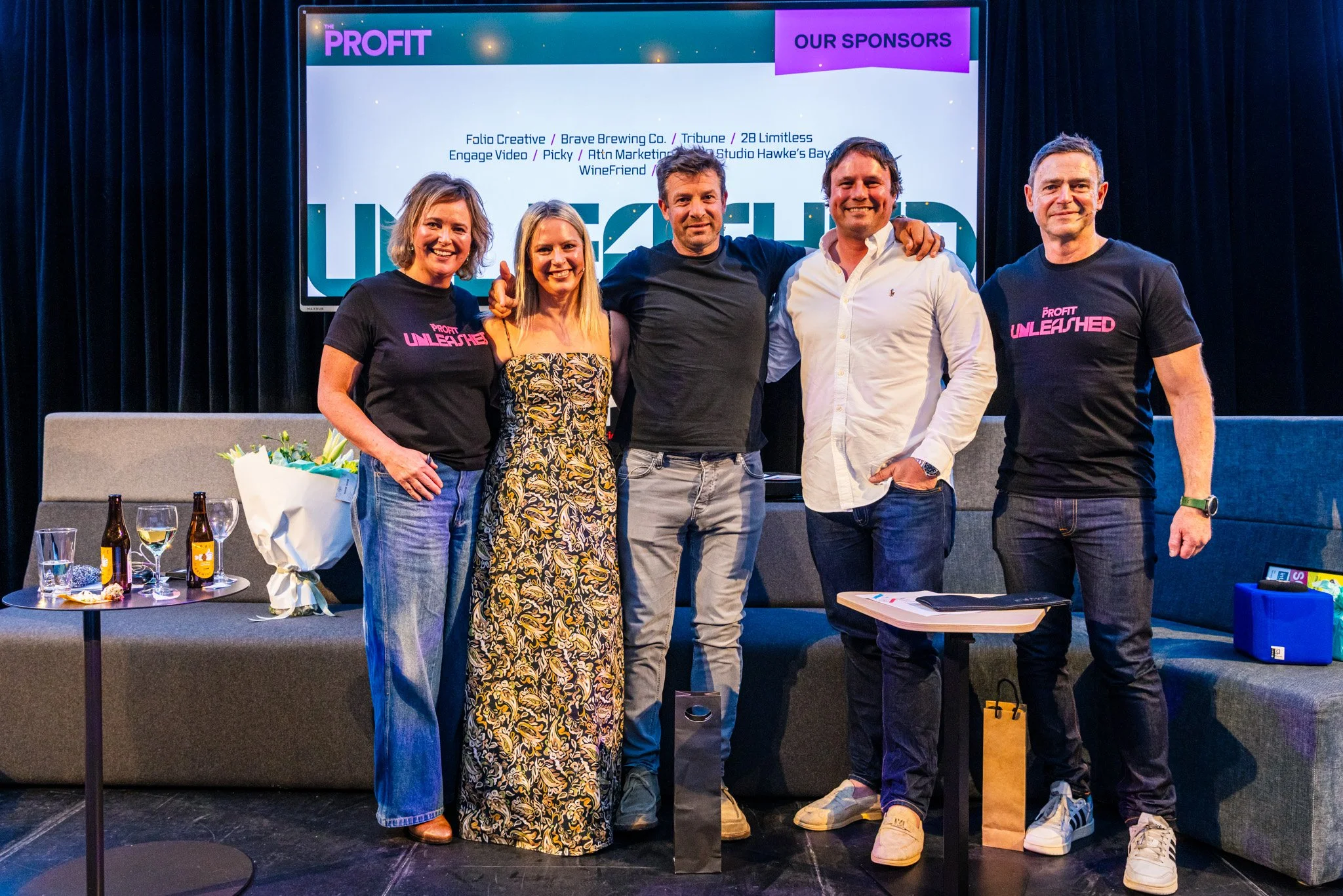Group of five smiling people standing together on a stage, with a large screen behind them displaying the word "PROFIT" and various sponsor names, in a dark room with black curtains. There are glasses, bottles, and a bouquet on a sofa and tables near