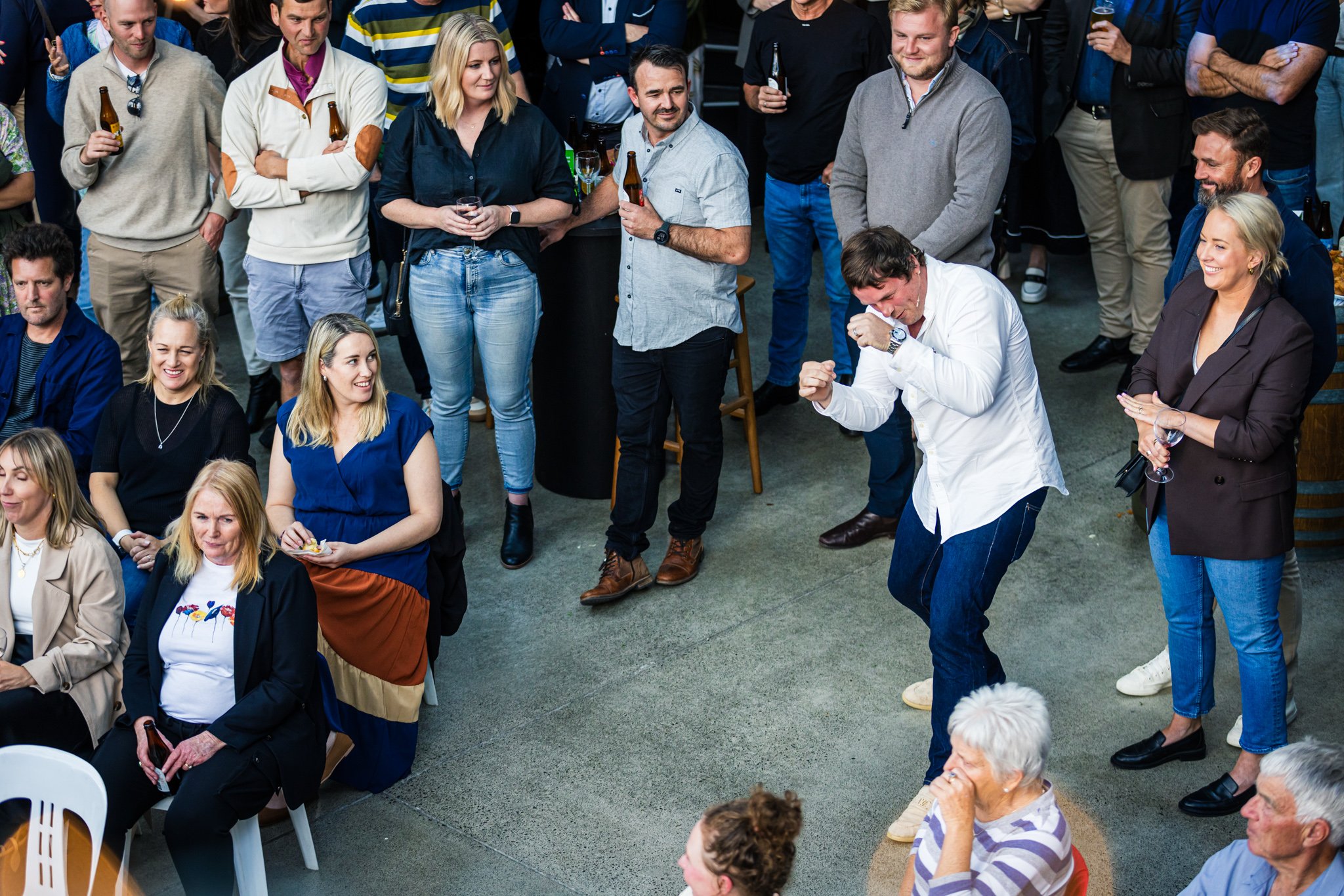 Group of people at an indoor event, some standing and some sitting, with one man dancing energetically in the center, surrounded by smiling and laughing guests holding drinks.