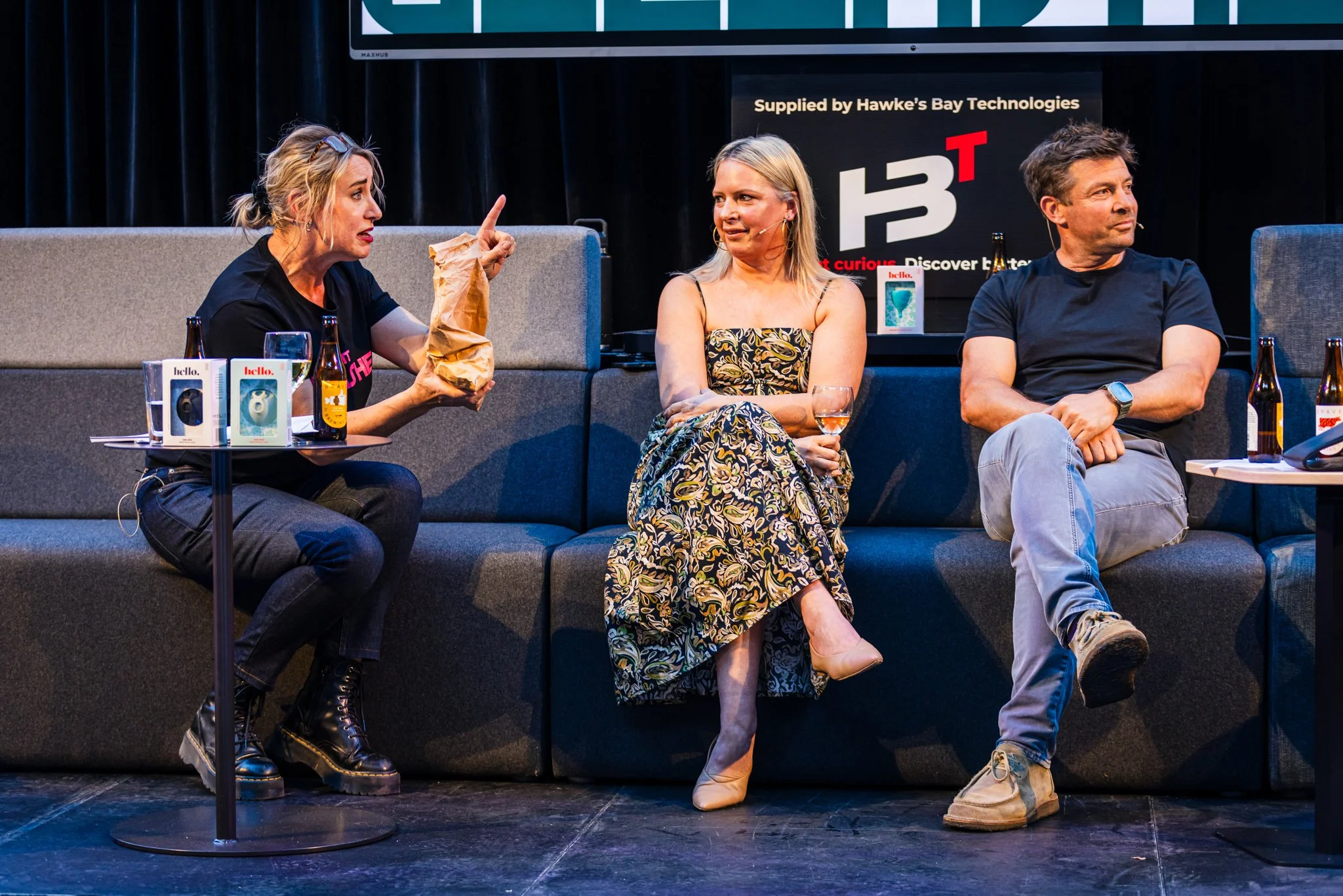Three people seated on a stage couch, participating in a discussion during a panel event. The woman on the left is talking and gesturing with her finger, the woman in the middle is holding a glass and listening, and the man on the right has his arms 