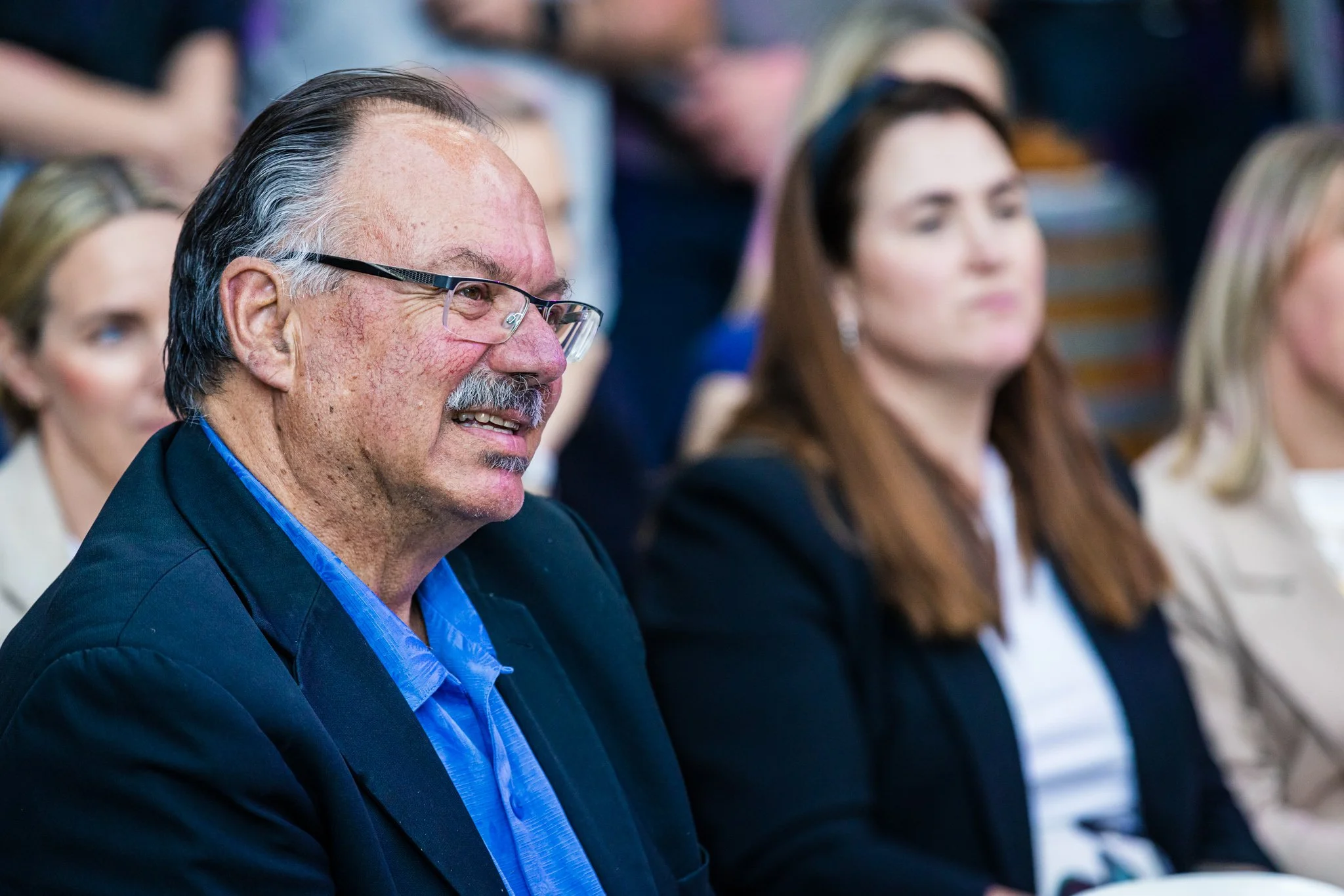 An older man with glasses, gray hair, and a mustache, smiling and wearing a dark blazer over a blue shirt, sitting in an audience with other people in the background.