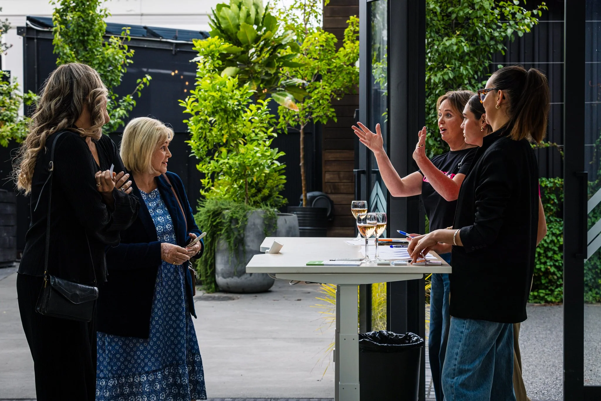 Four women standing outside near a reception table with glasses of wine, engaged in a conversation. One woman is gesturing as she speaks, while two women listen attentively.