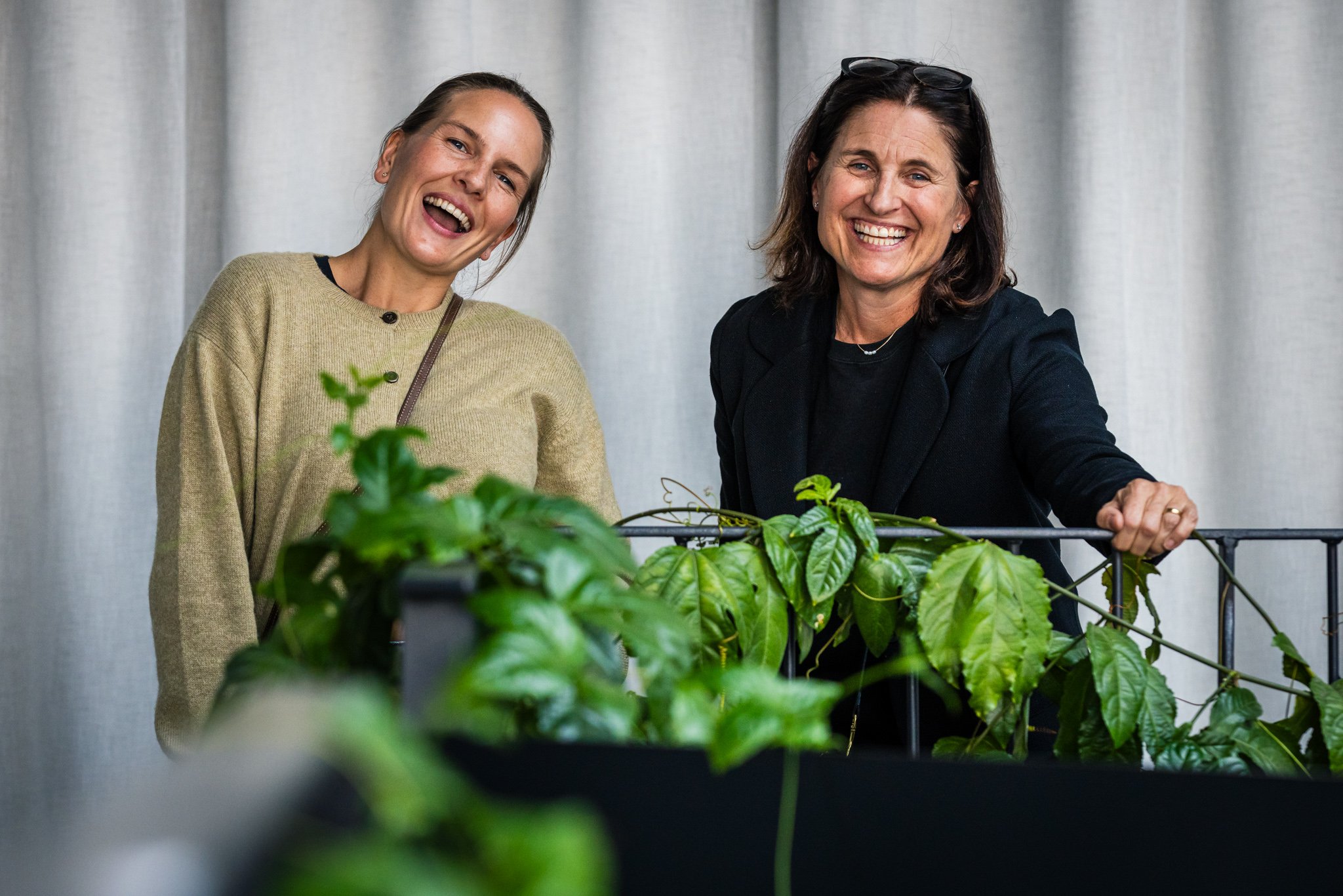 Two women smiling and interacting with green plants in a bright indoor setting.