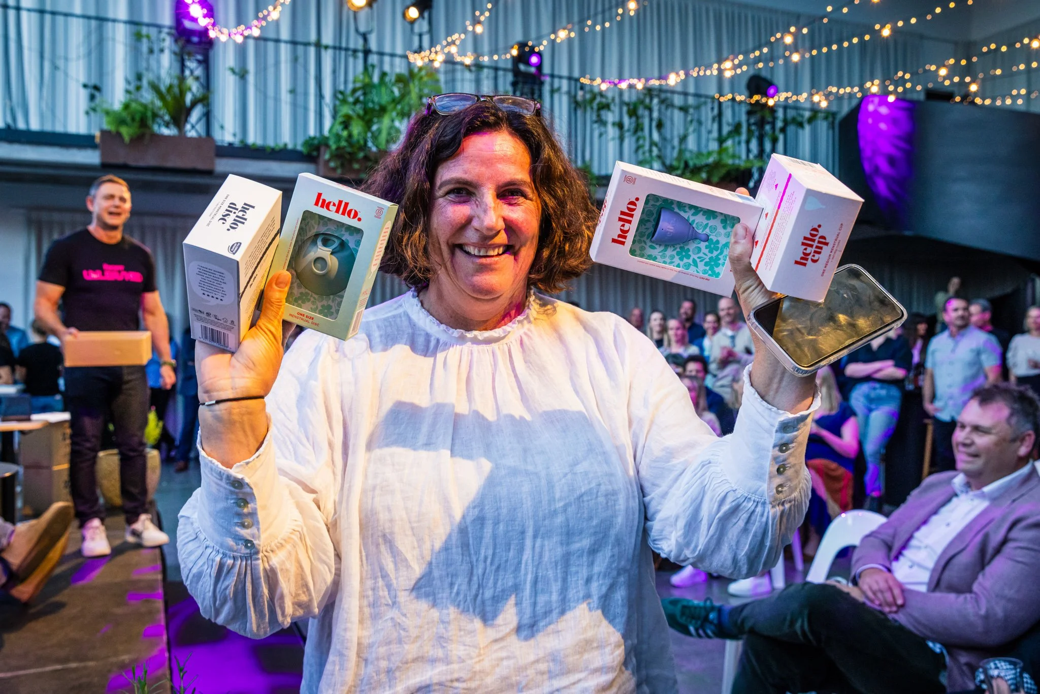 A woman smiling and holding up multiple boxed products at an indoor event with string lights and crowd in background.
