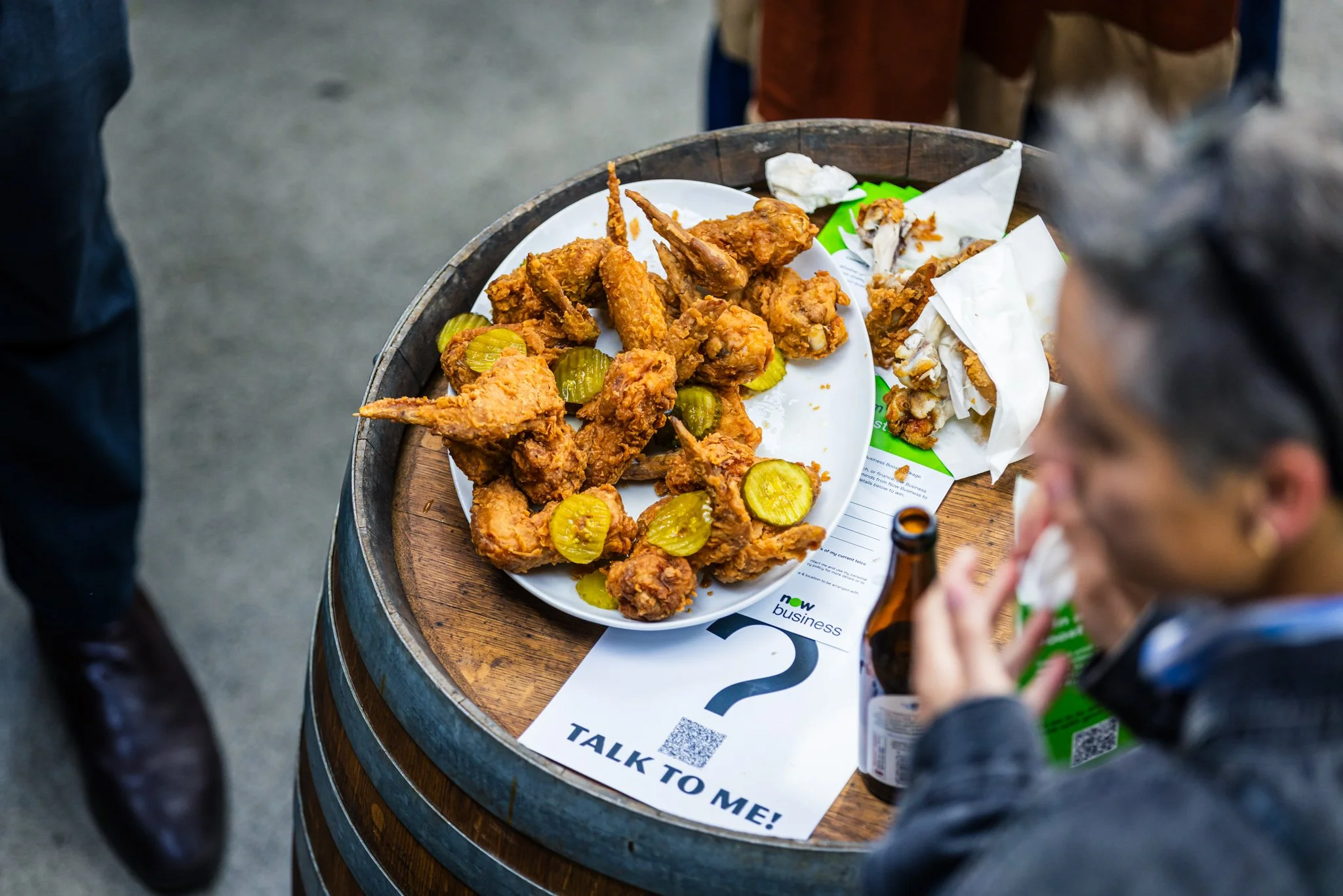 Plate of fried chicken wings and pickles on a barrel-shaped table, with a person holding a beer bottle and talking, at an outdoor event.