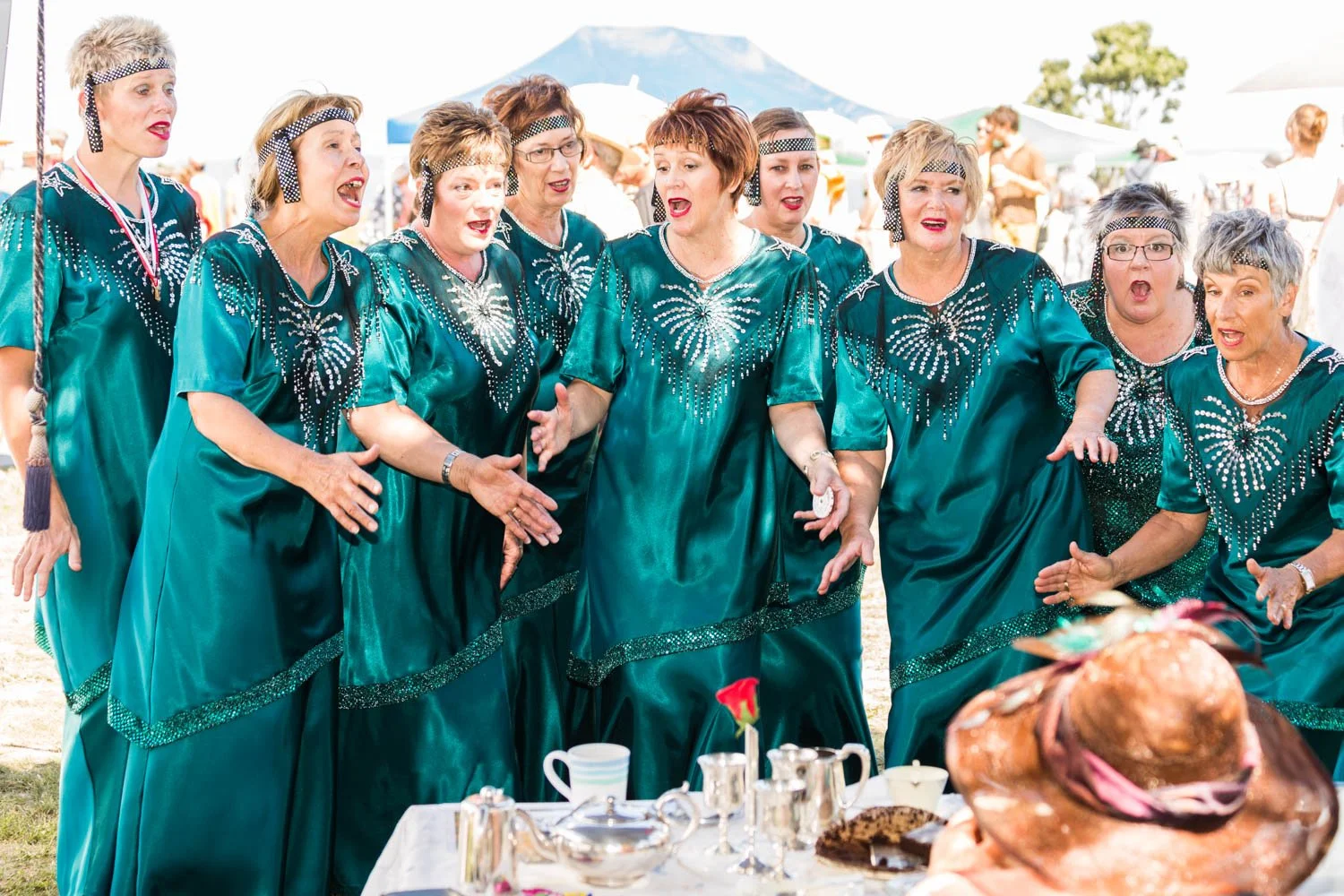 A group of women dressed in matching teal, sparkly dresses singing and performing outdoors at a gathering, with a table of food and drinks in front.