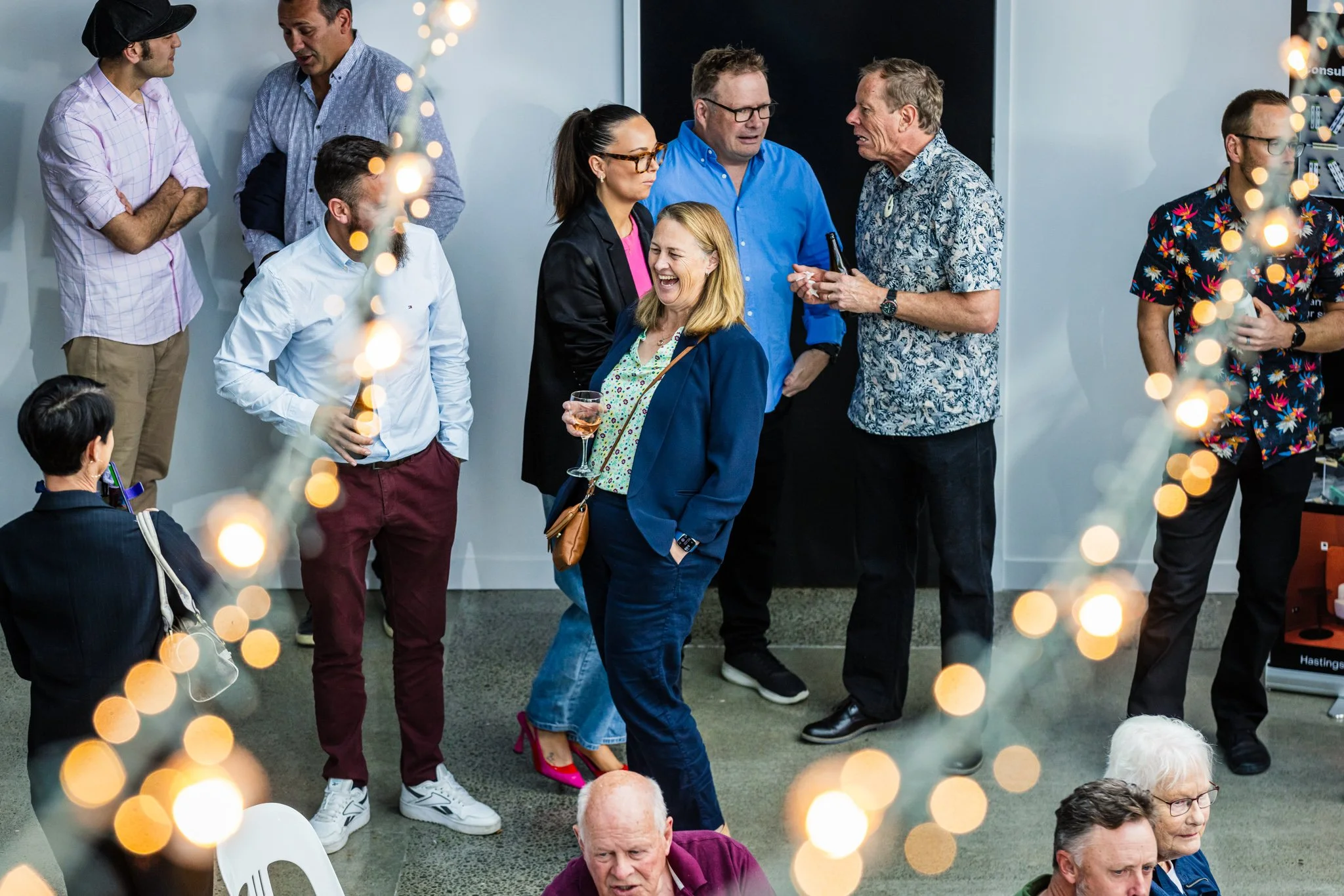 People socializing at a party with string lights, some standing and conversing, one woman laughing with a drink in hand, diverse group in casual attire.