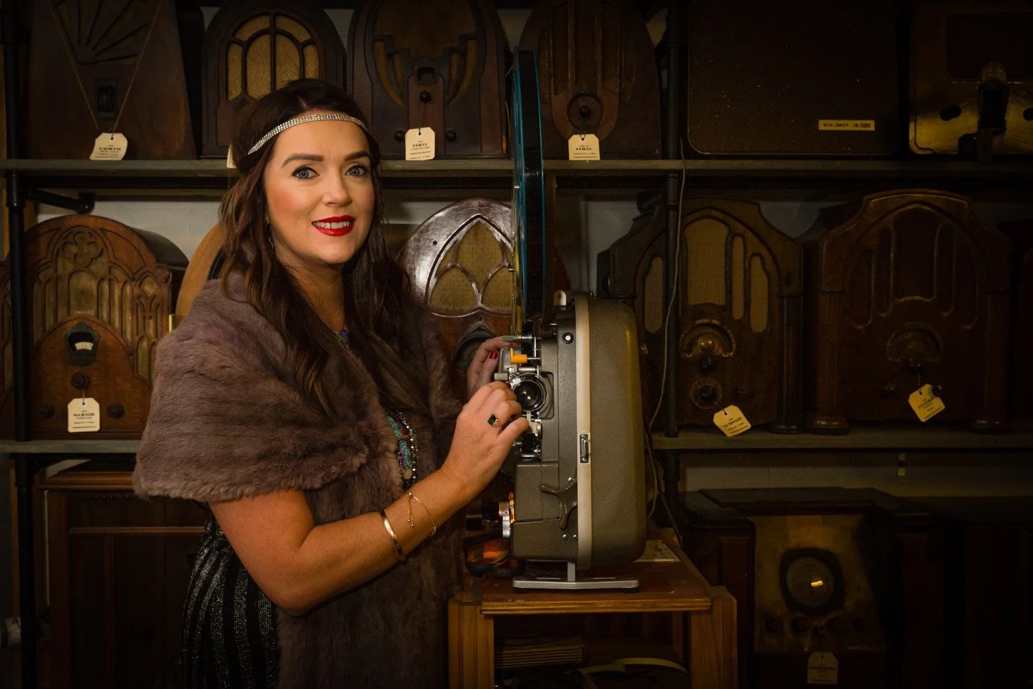 A woman with dark brown hair, red lipstick, and a sparkle headband using a vintage film camera in a room with shelves of old radios.