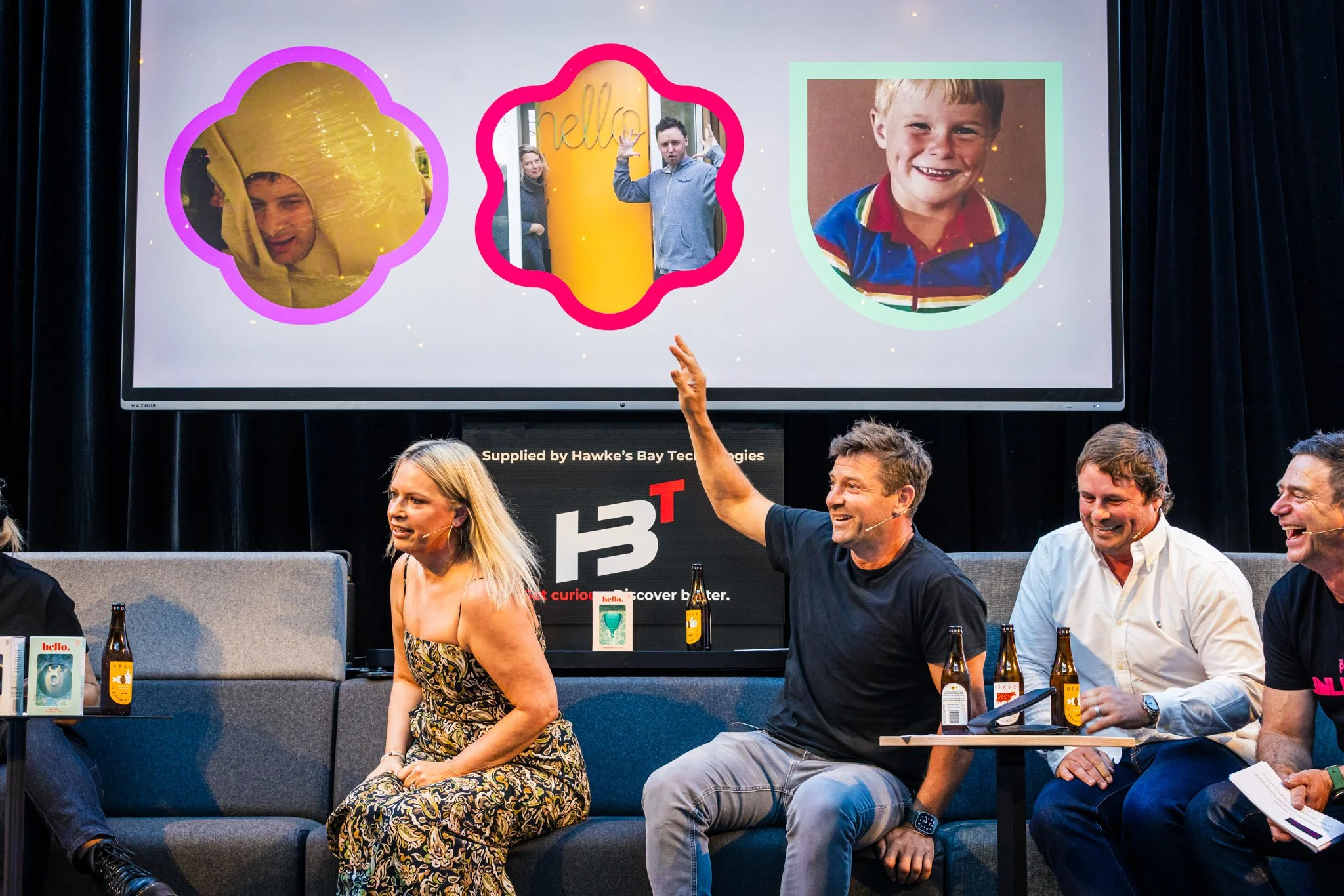 A group of five people seated on a stage, participating in a discussion or panel. There are bottles of drinks on the table in front of them. A man in the center is raising his hand, smiling. Behind them is a large screen displaying three photos: a ch