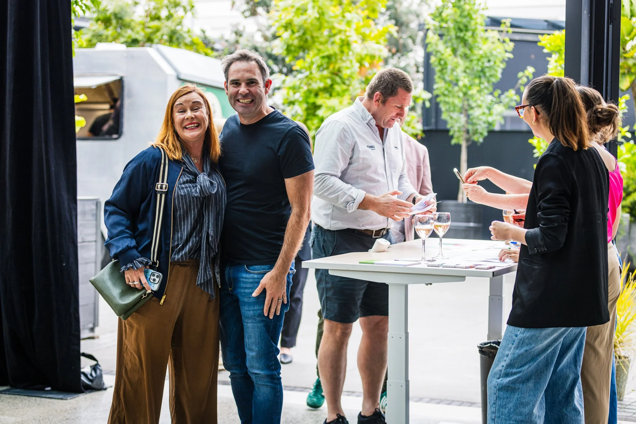 A group of people at an outdoor event, with two people smiling at the camera in the foreground and others interacting at a registration or check-in table in the background, surrounded by green trees.