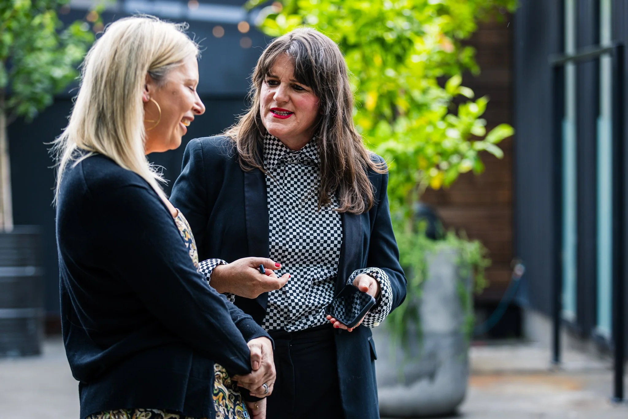 Two women engaging in a conversation outdoors, with greenery and modern architecture in the background.