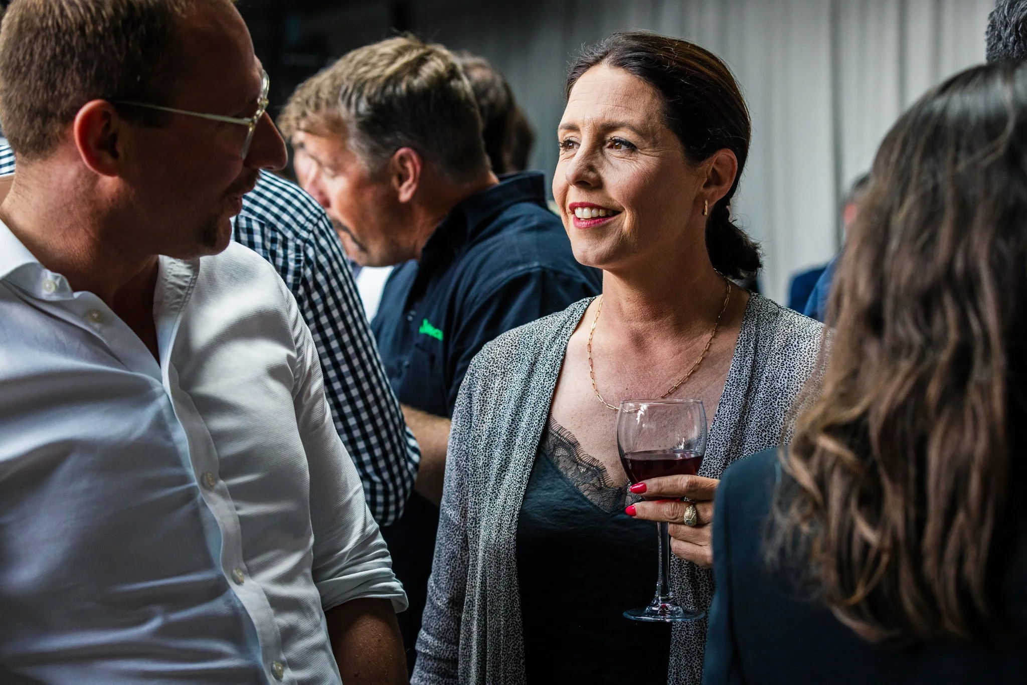 A woman holding a glass of red wine talking to a man at a social gathering, with other people in the background.
