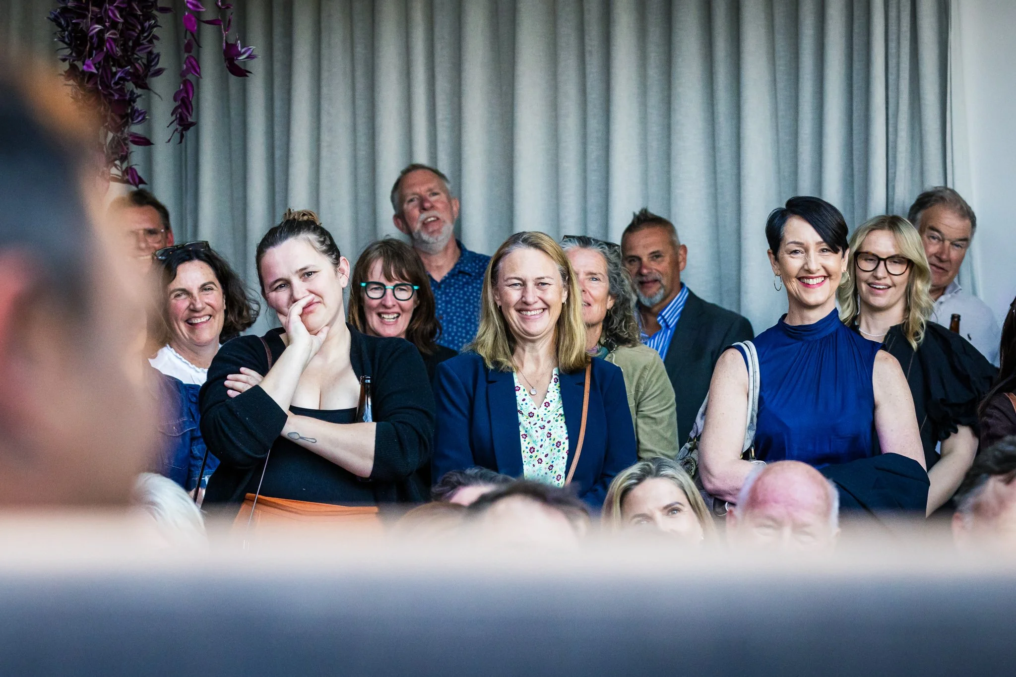Group of smiling people, mostly women, attending an indoor event with gray curtains in the background.