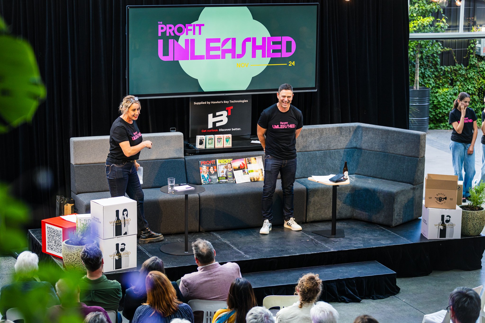 A presentation event with two speakers on stage, one woman and one man, standing in front of a large screen that reads "Profit Unleashed." The woman is on the left, holding papers and smiling, while the man is on the right, also smiling. There are ta