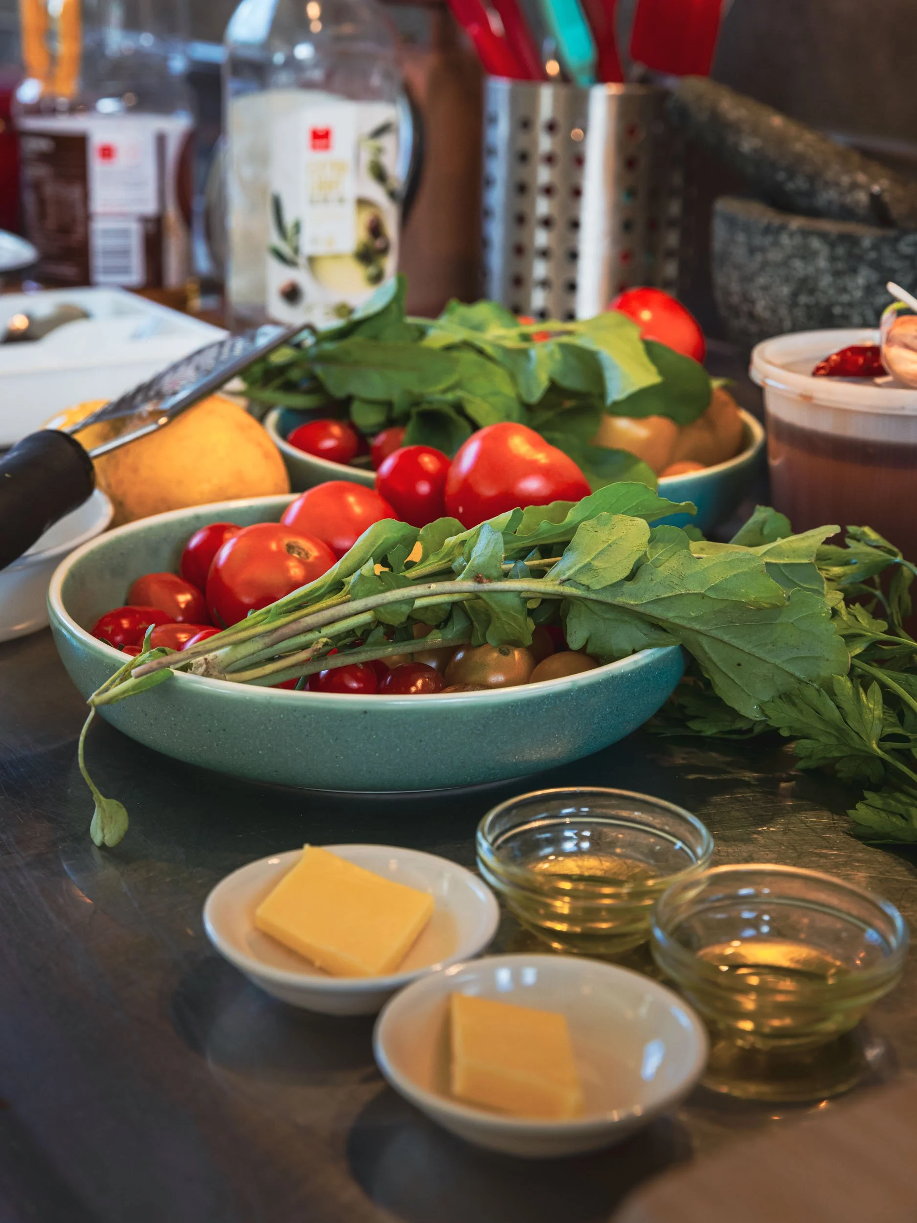 Bowls of cherry tomatoes, fresh greens, and herbs on a kitchen counter, along with small dishes of butter and olive oil, preparing for cooking.