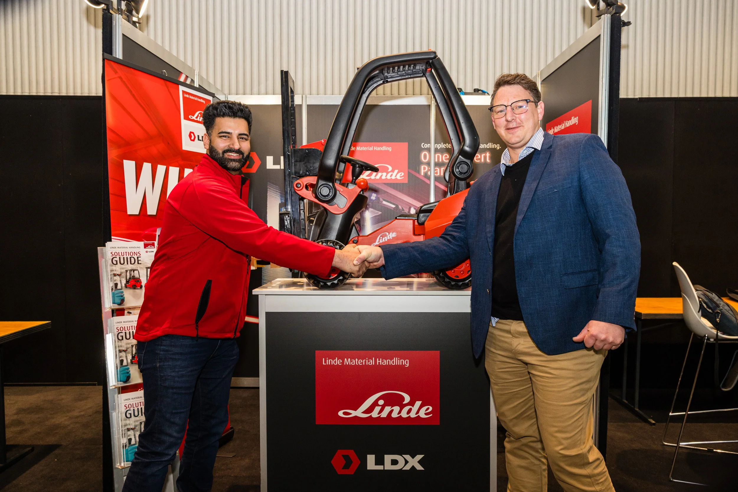 Two men in business attire shaking hands at an exhibition booth with a red and black display for Linde Material Handling and LDX, showcasing a small industrial forklift.