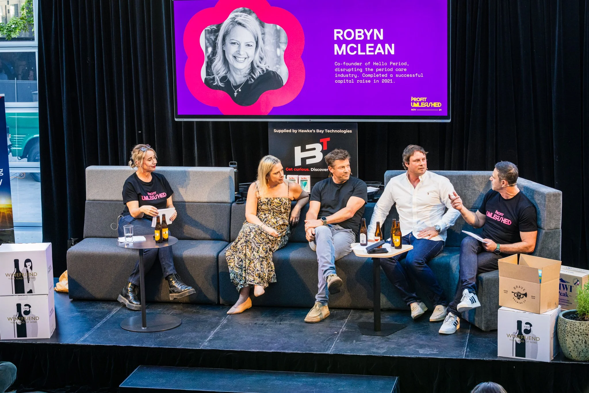 A panel discussion with five people seated on a stage, three men and two women. One woman is wearing a buffet dress, and the other is in a black t-shirt with pink text. They are seated in front of a large screen displaying a woman’s photo and bio, al