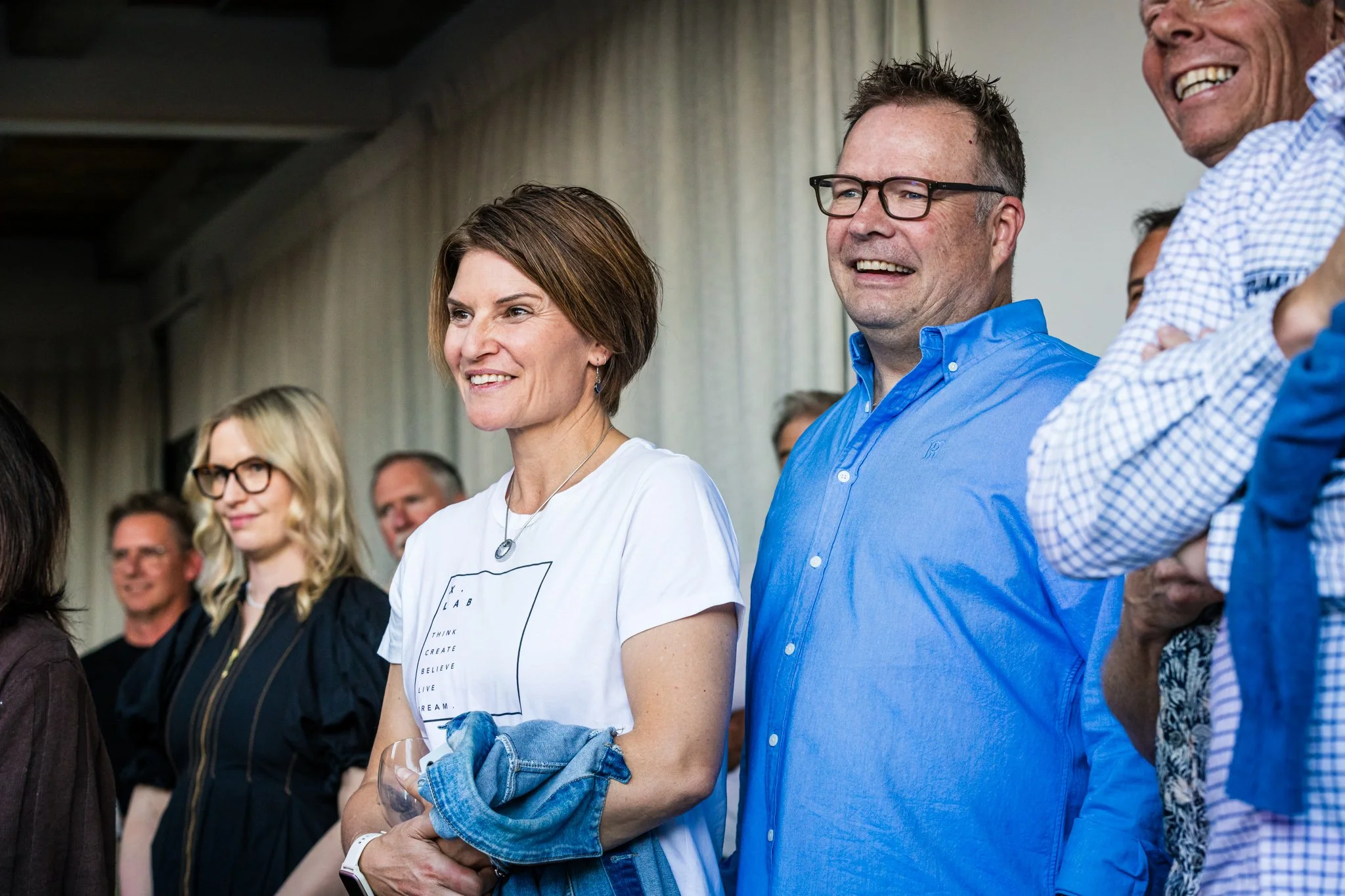 People standing together in a room, smiling, and paying attention to something outside the frame, with a woman in a white shirt and glasses and a man in a blue shirt in the foreground.
