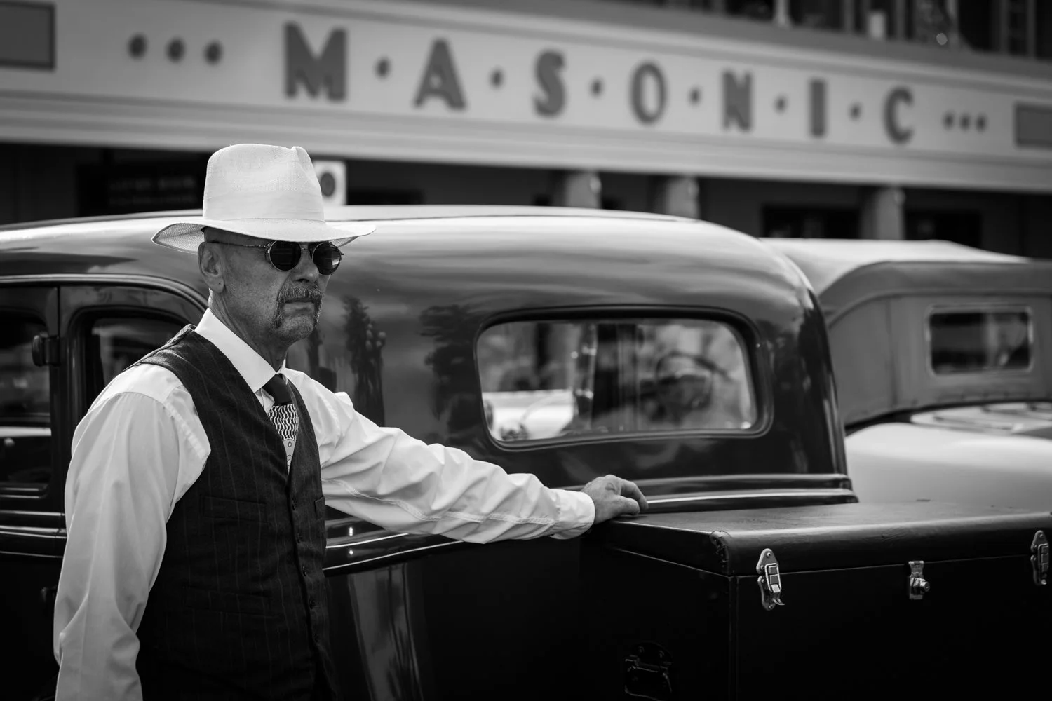 A man dressed in vintage clothing, wearing sunglasses and a light-colored fedora hat, standing next to an old-fashioned truck in front of a Masonic theater building.