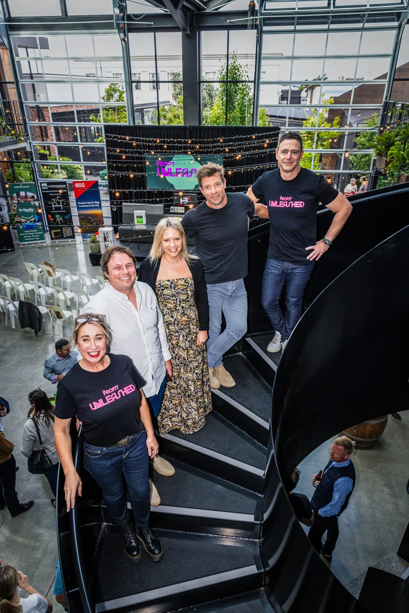 Group of five people standing on a black spiral staircase inside a modern, glass-walled building with string lights and a large screen displaying 'Profit Unleashed' in the background. Two women are wearing black T-shirts with 'Profit Unleashed' writt