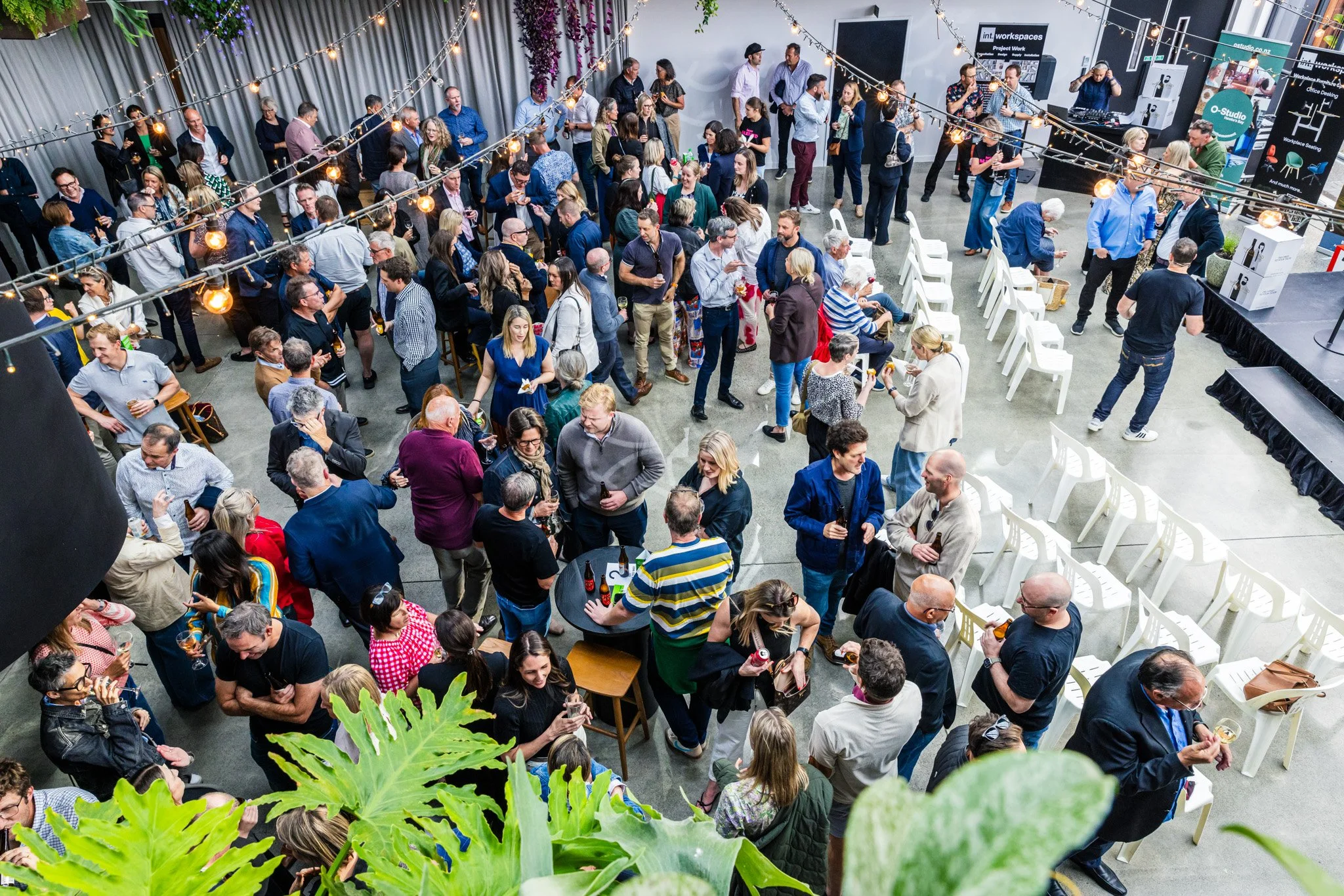 A large indoor networking event with people socializing, standing, and sitting around high tables, some holding drinks. Decor includes string lights and plants, with booths and screens in the background.