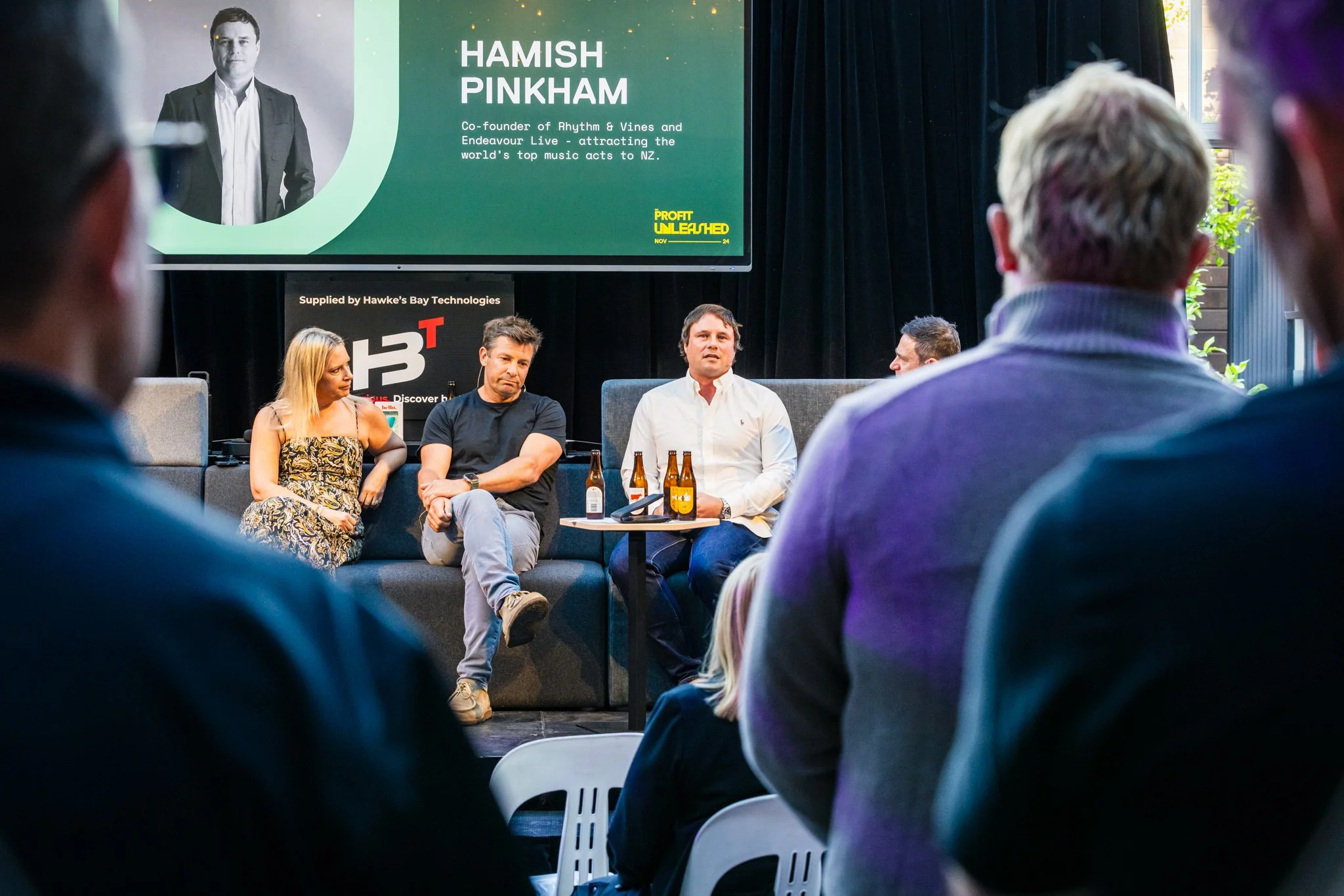 Panel discussion at an event featuring Hamish Pinkham, with a large screen displaying his name and credentials, and seven people in the audience facing the speakers.