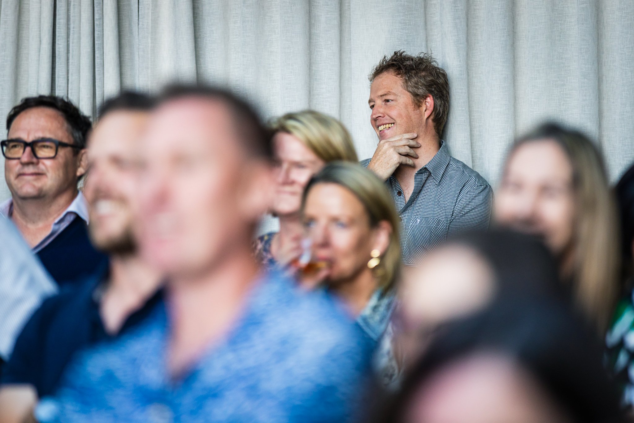 A group of people attending an indoor event, with a man in a checkered shirt smiling and touching his chin, seated against a grey curtain backdrop.