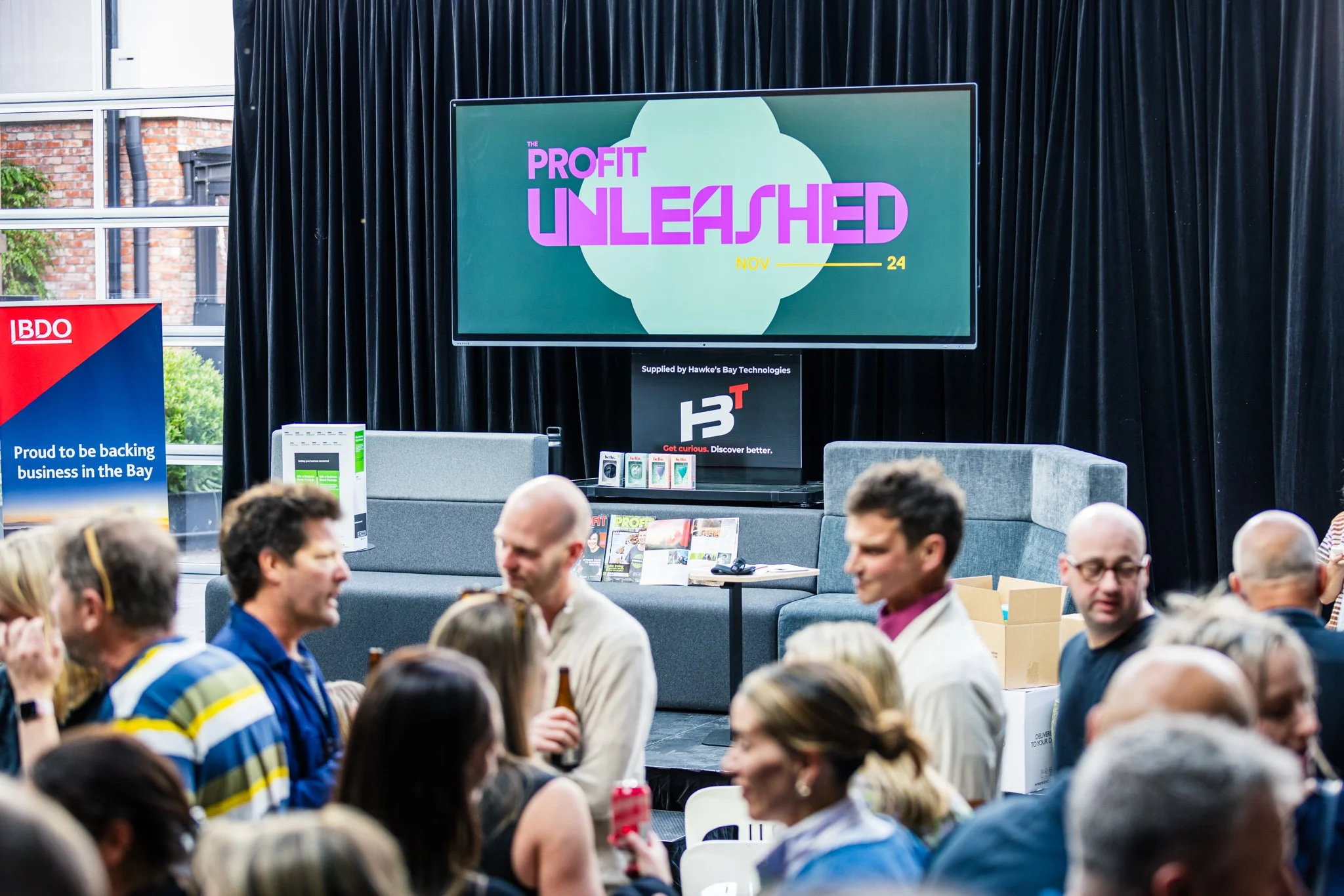 Conference room with audience attending an event titled 'Profit Unleashed' on a large screen, featuring a stage with chairs, tables, and promotional signage.