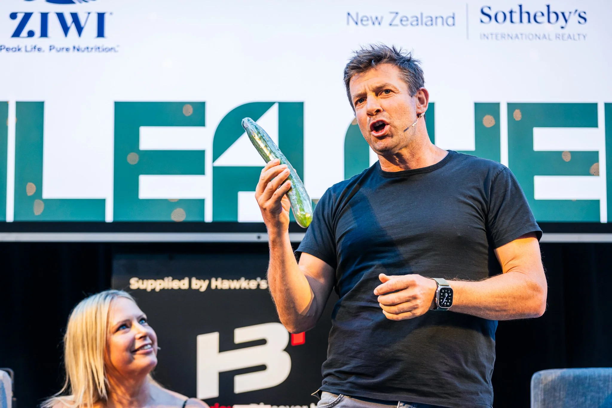 A man in a black t-shirt holding a pickle and speaking on stage, with a woman smiling at him and a large sign in the background.