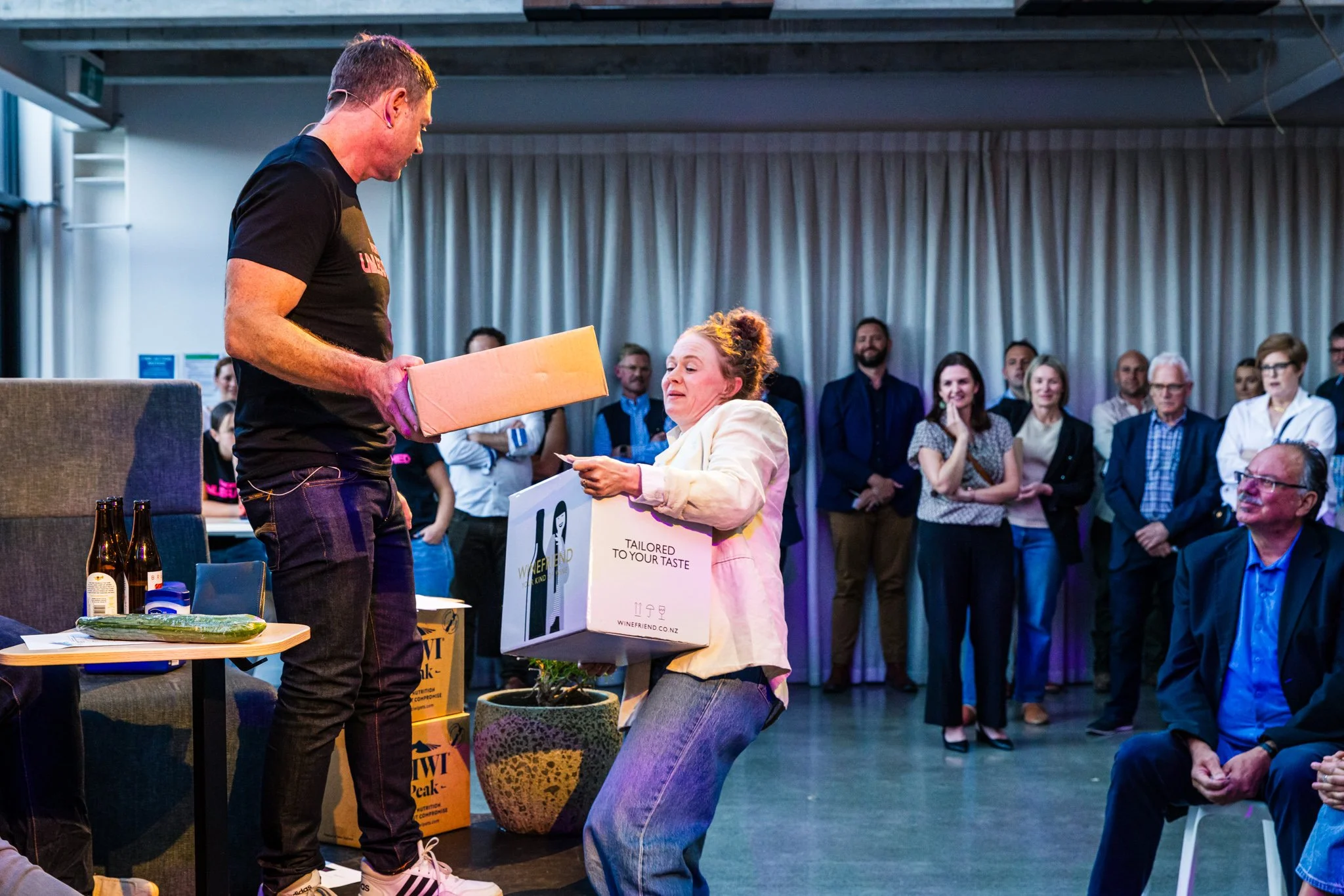 A woman receiving a gift box from a man during an indoor event, with a group of people watching in the background.