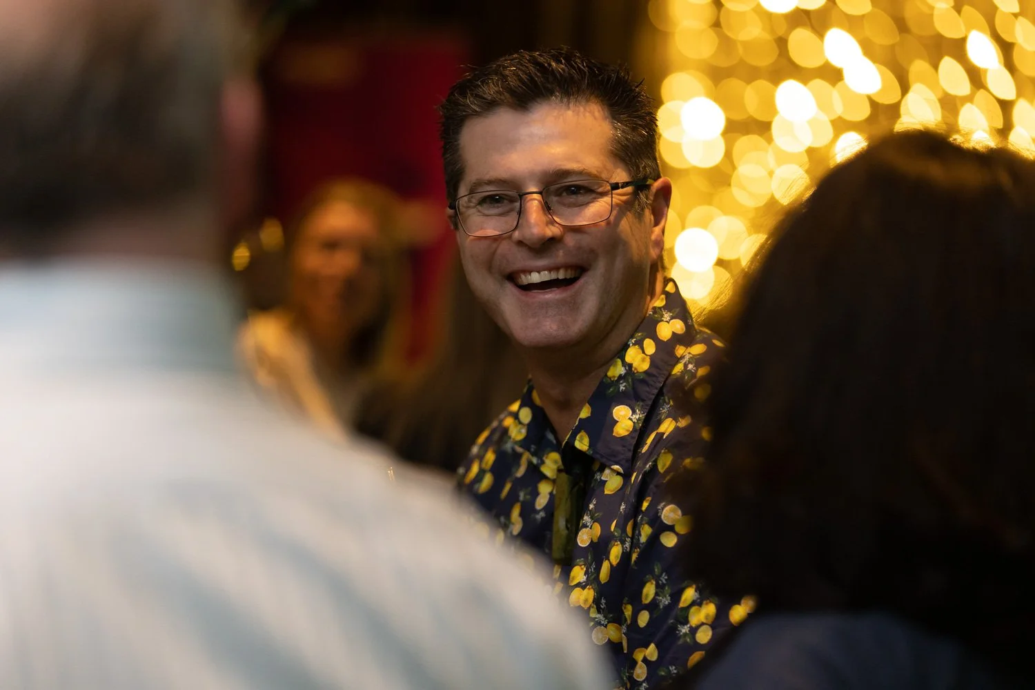 A man with glasses smiling and talking to people at a gathering, with warm-colored bokeh lights in the background.