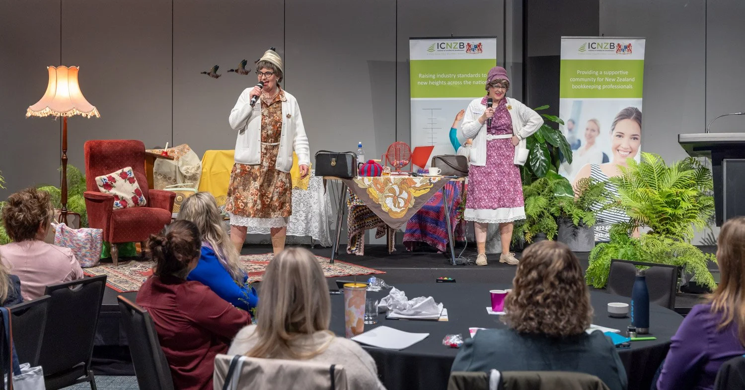Two women are on a stage, speaking into microphones at an event. They are dressed in vintage-style clothing and period costumes. The stage has a table with colorful knitting and crocheting items, and a decorative armchair. There are potted plants and