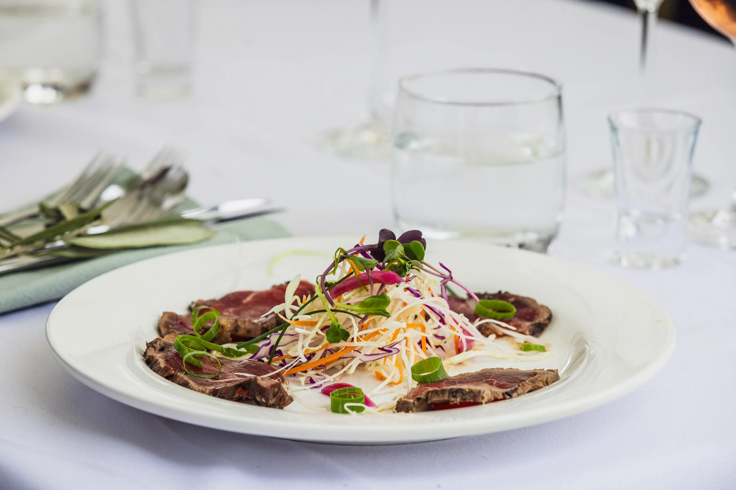 A plate of sliced steak with a side of coleslaw salad topped with microgreens on a table set for a meal.