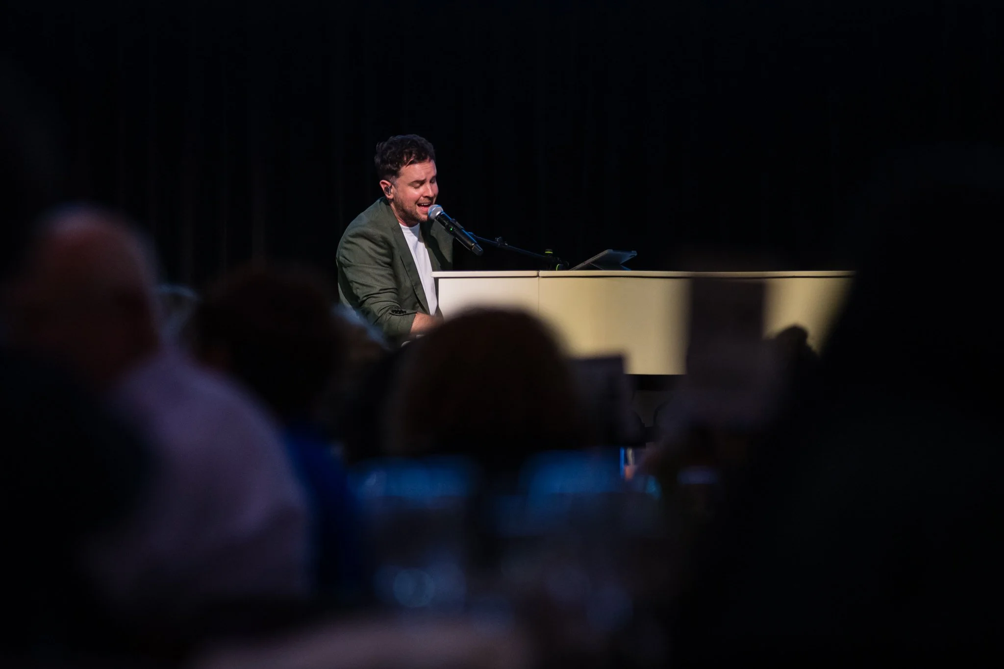Man playing piano and singing into microphone on stage at a concert or event, with audience members in foreground.