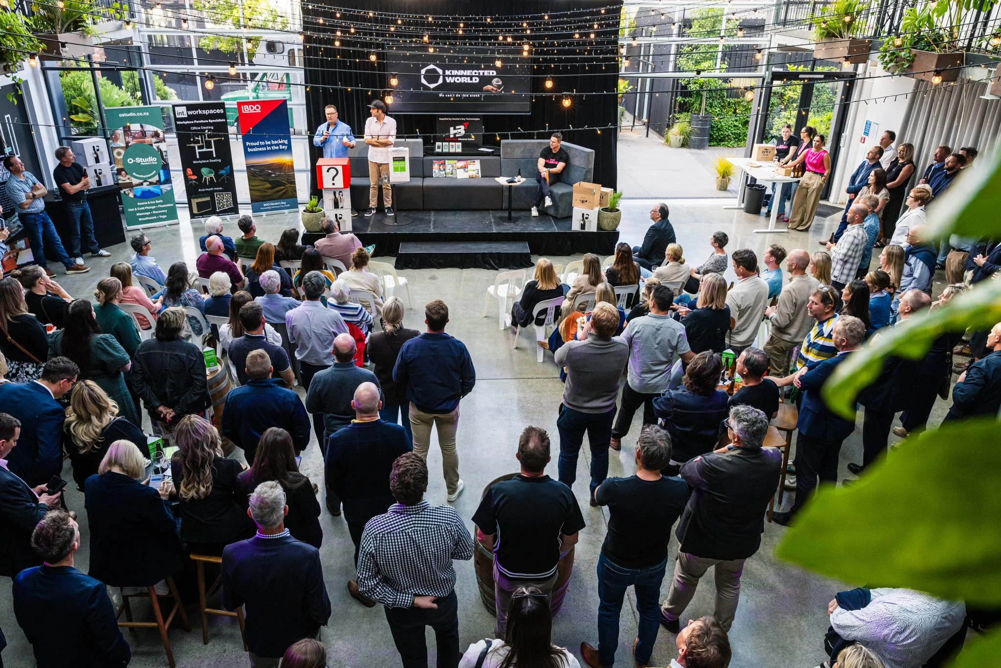 Indoor event with a speaker and audience, with banners and decorations, high ceiling with string lights, and people standing and sitting while listening.