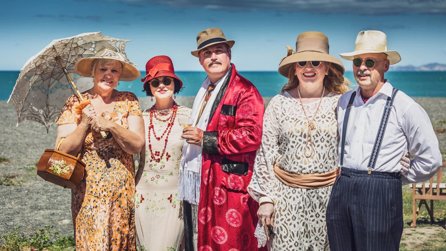 Six adults dressed in vintage beach attire standing on a beach with the ocean in the background.