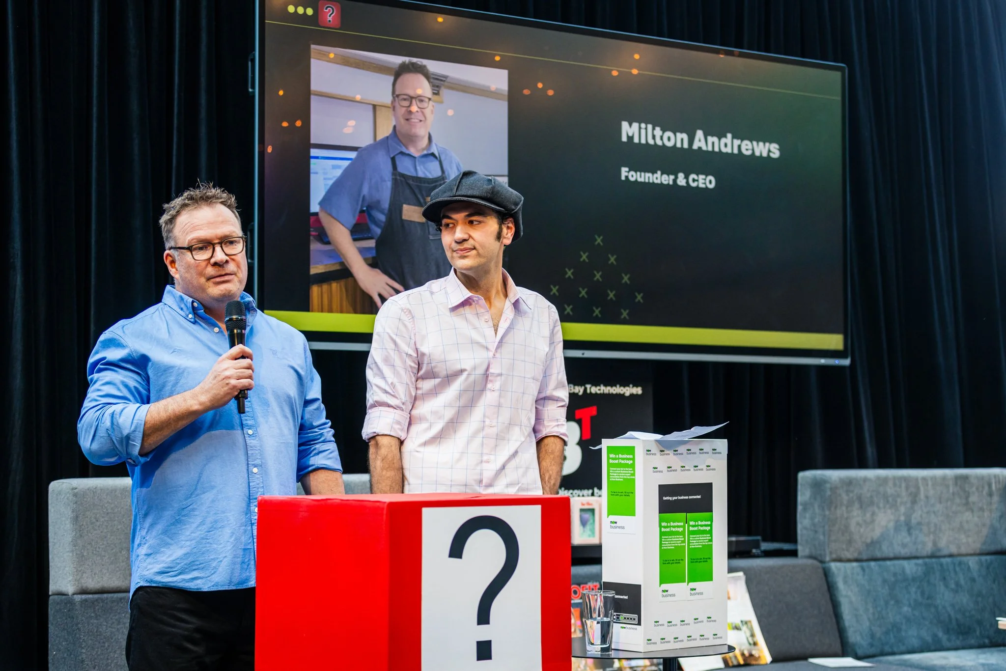 Two men standing on a stage, one holding a microphone. Behind them, a large screen displays a photo and text reading 'Milton Andrews, Founder & CEO'. There is a large red block with a question mark on it, with a white box containing pamphlets or prin