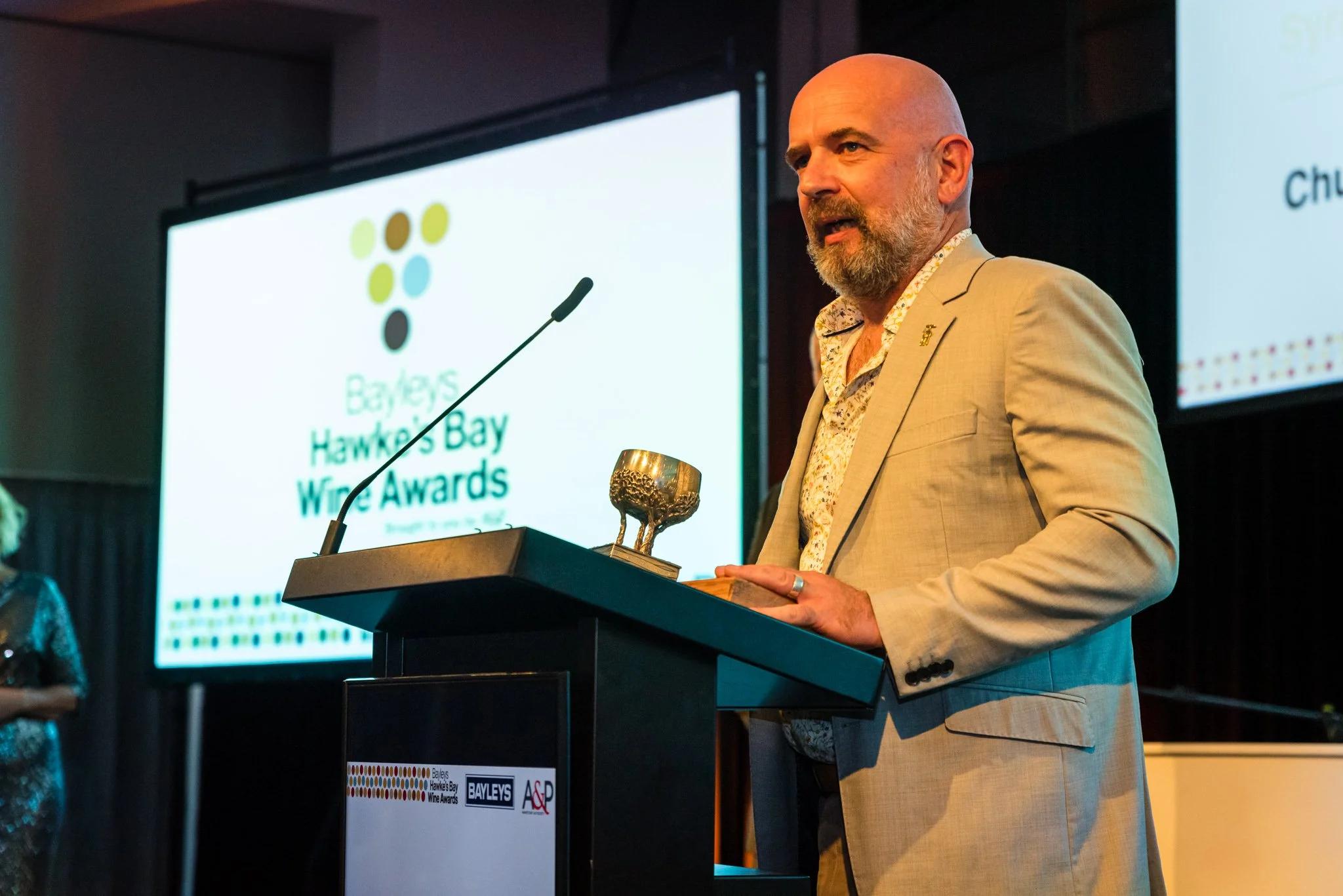 A man in a beige suit speaking at a podium during the Bayleys Hawkes Bay Wine Awards, with a large screen behind displaying the event logo and name.
