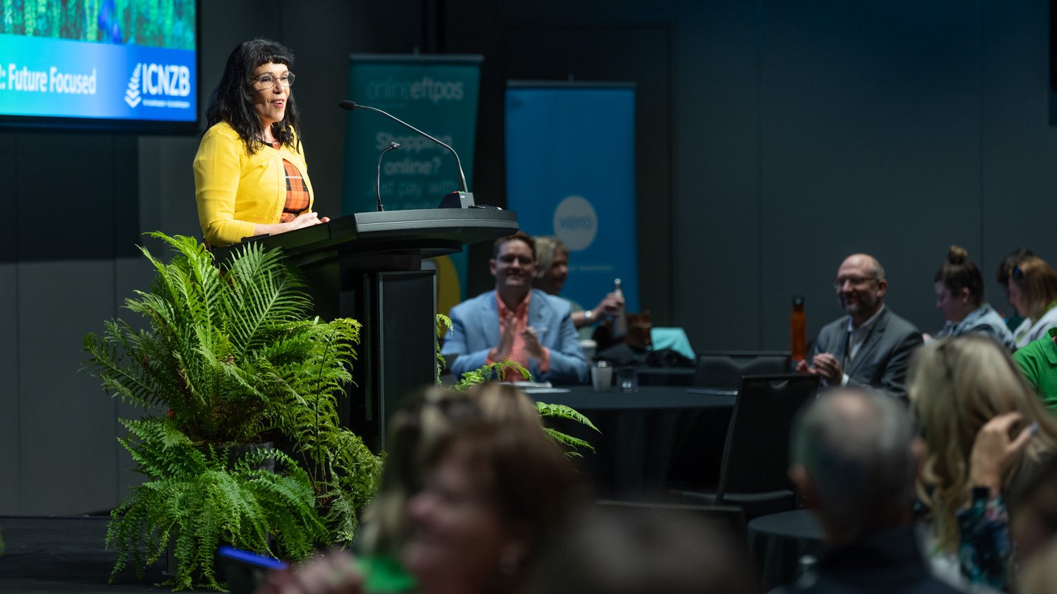 Woman in yellow jacket speaking at a podium with plants in front, audience listening and clapping in the background at conference.