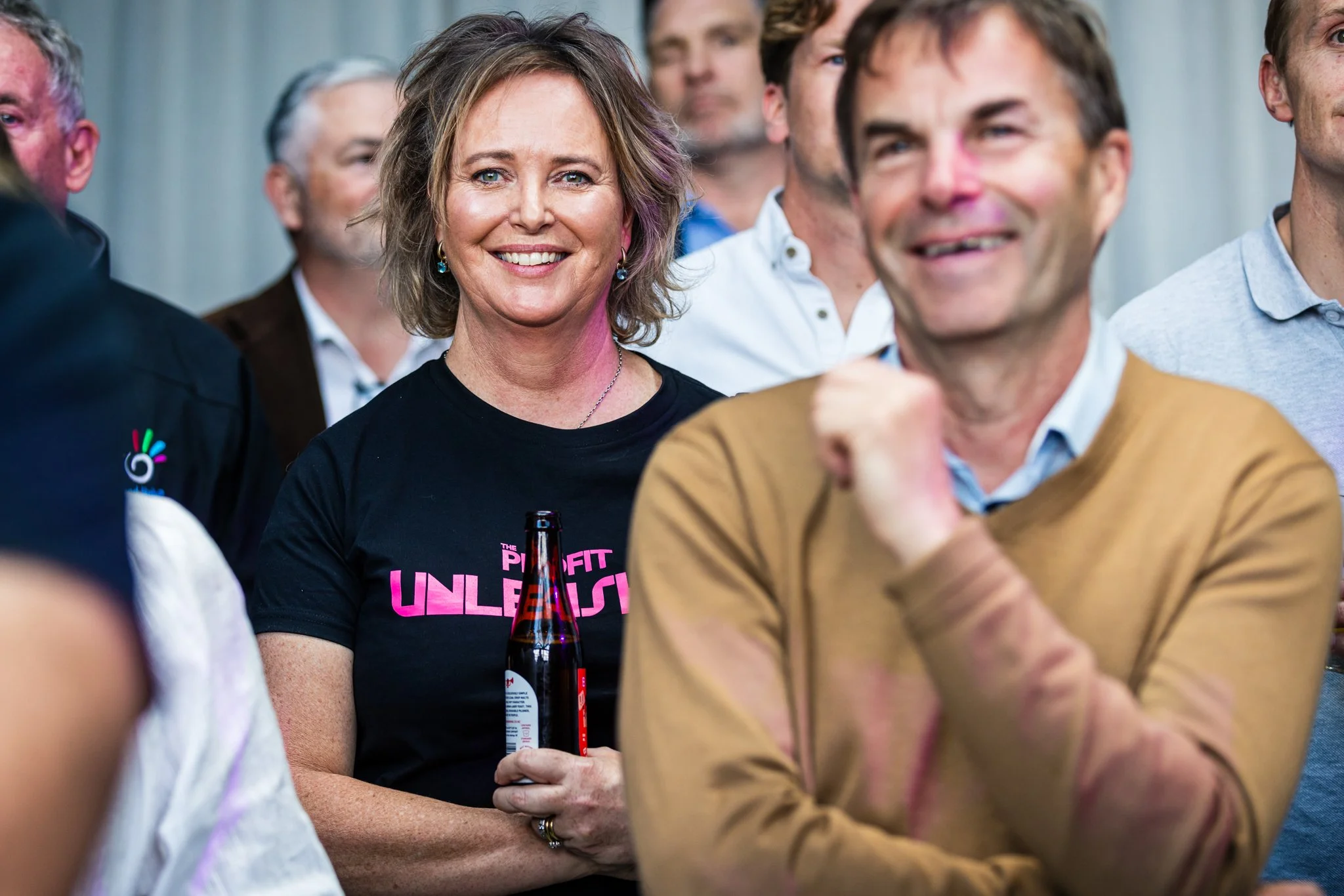 A group of people at an event, with a woman in the center smiling and holding a beer bottle, wearing a black T-shirt with pink text that says 'The Profit UNLEASHED'.