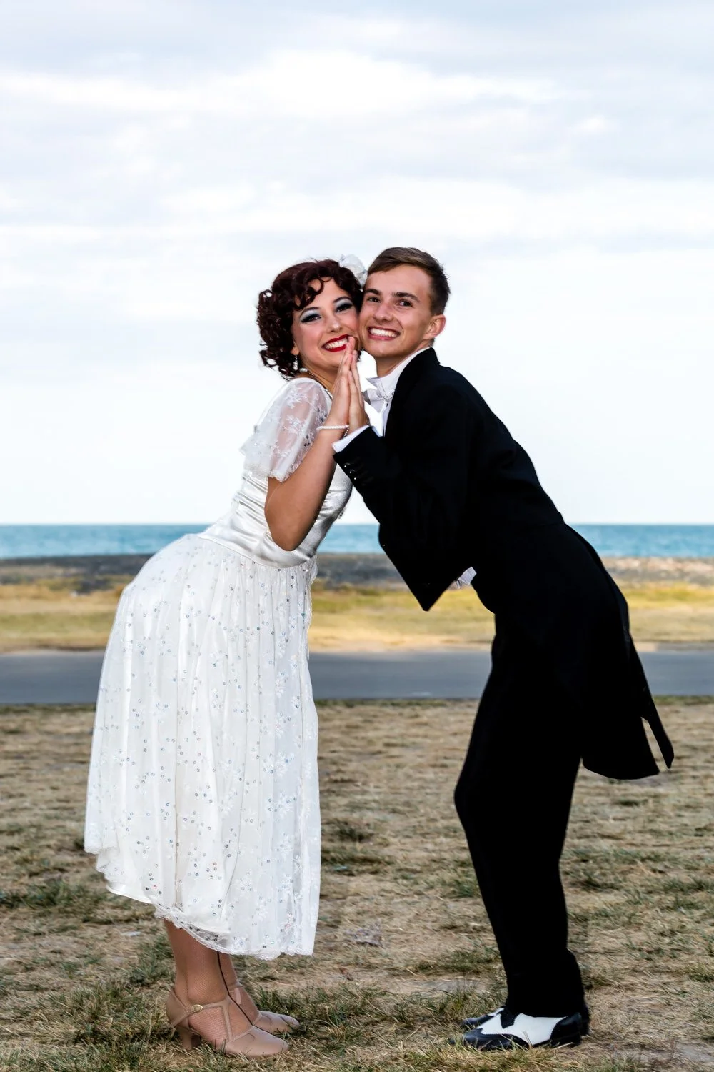 A couple dressed in vintage clothing, the woman in a white dress and the man in a black tuxedo, on a grassy area near the ocean, smiling and leaning their hands together.