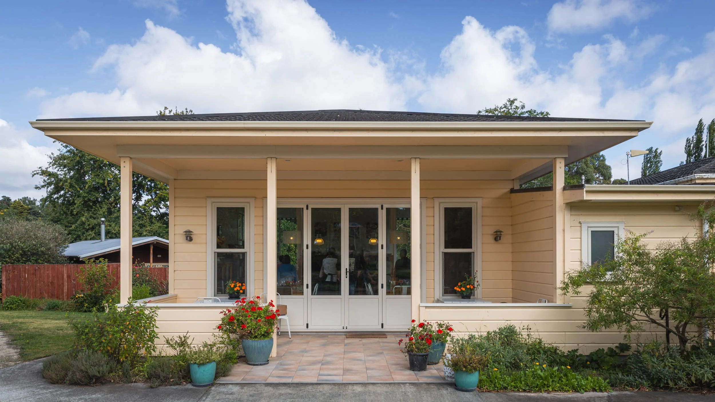 Front view of a house with a porch, potted plants, and a garden in the foreground under a partly cloudy sky.