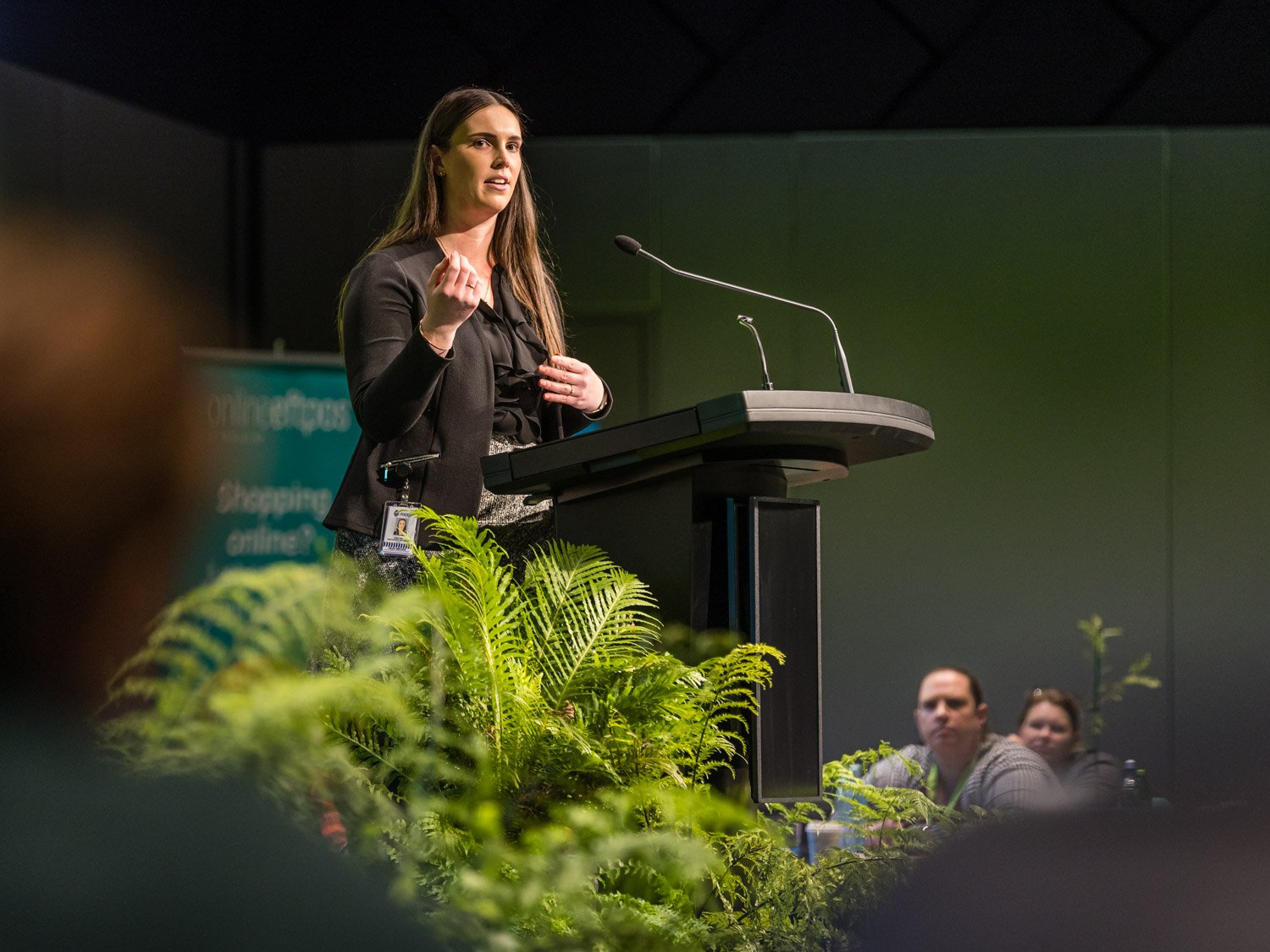 A woman speaking at a podium during a conference or seminar, with a badge on her waist, green plants in the foreground, and an audience seated behind her.
