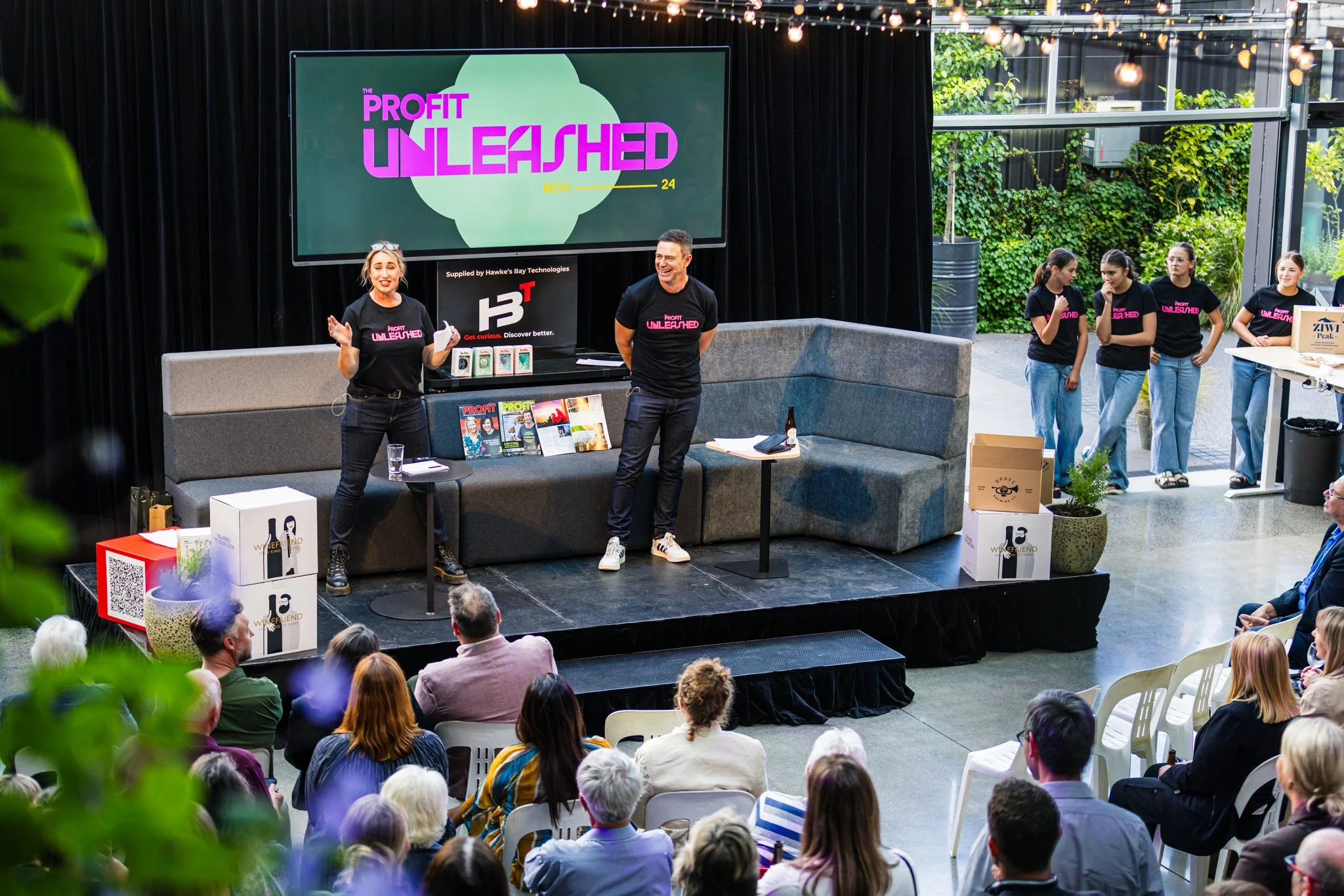 A conference or seminar event with a presenter on stage in front of a seated audience. The stage has a large screen displaying 'Profit Unleashed' with smaller text underneath. The presenter, a woman, is speaking with a man standing nearby, both weari