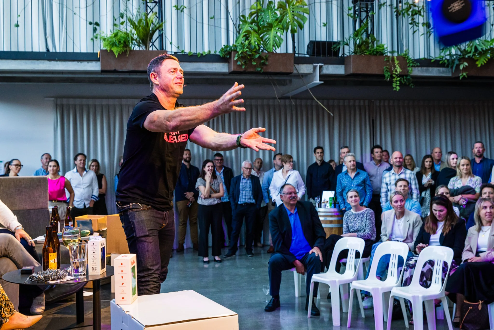 A man in a black t-shirt standing and gesturing with hands as he speaks to an audience in a large room. Behind him, a group of people is watching, some seated and some standing, with a curtain and plants in the background.