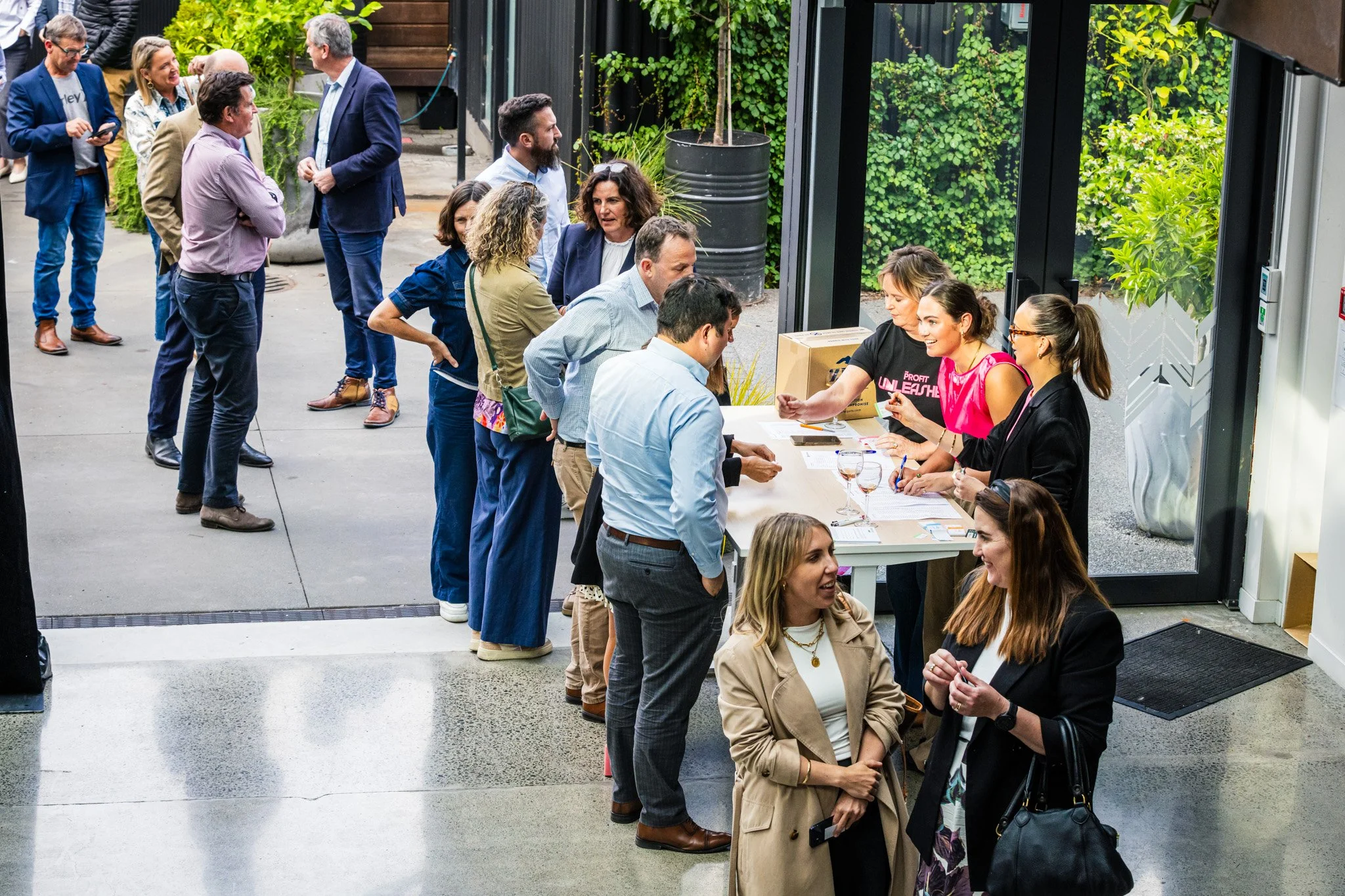 People lining up at a registration table during an event in a modern indoor space with glass doors and greenery outside.