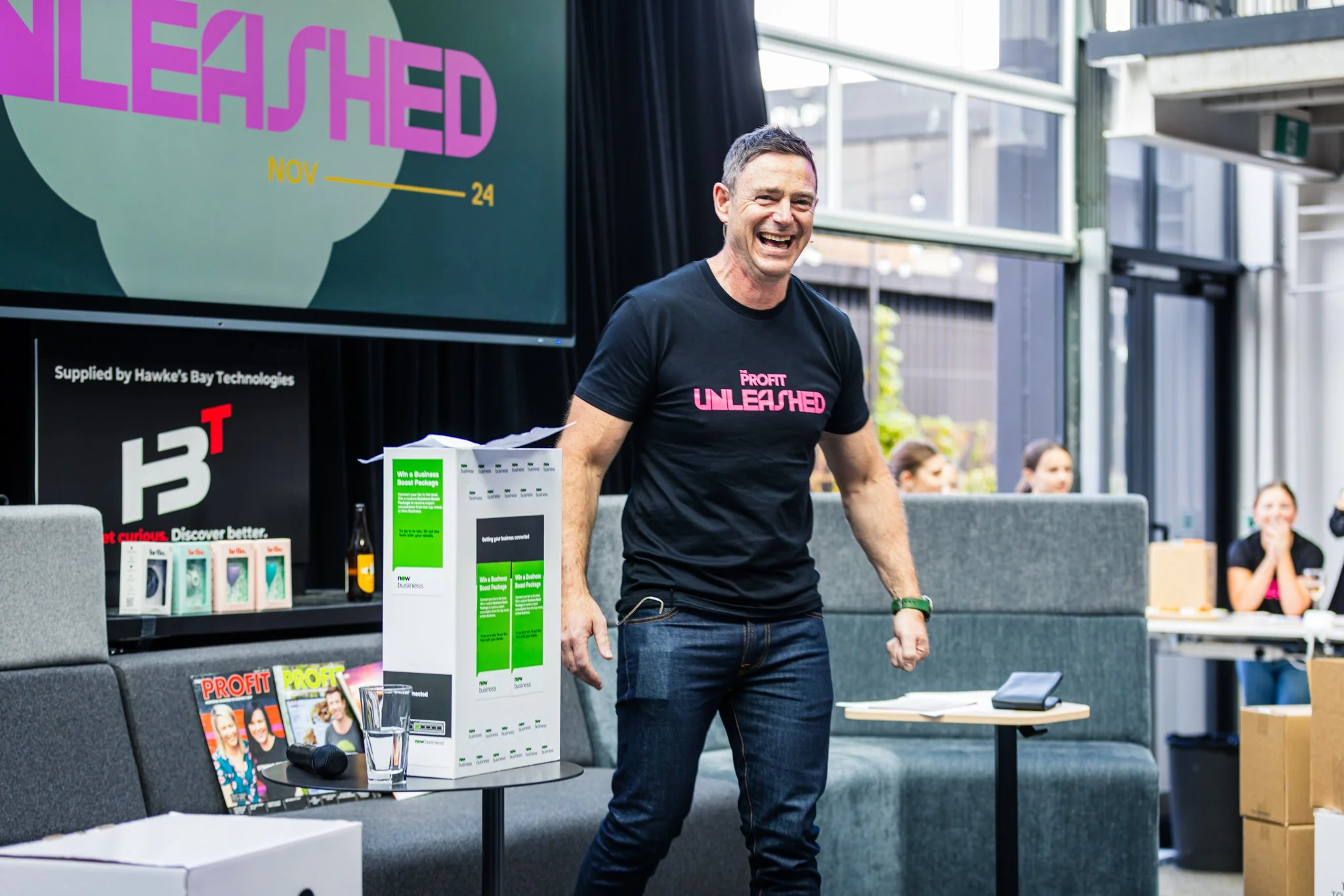 A man is smiling and standing in front of a presentation screen that says "Profit Unleashed". He is wearing a black t-shirt with the same text and has a small microphone clipped to his sleeve. The setting appears to be an indoor event space with tabl
