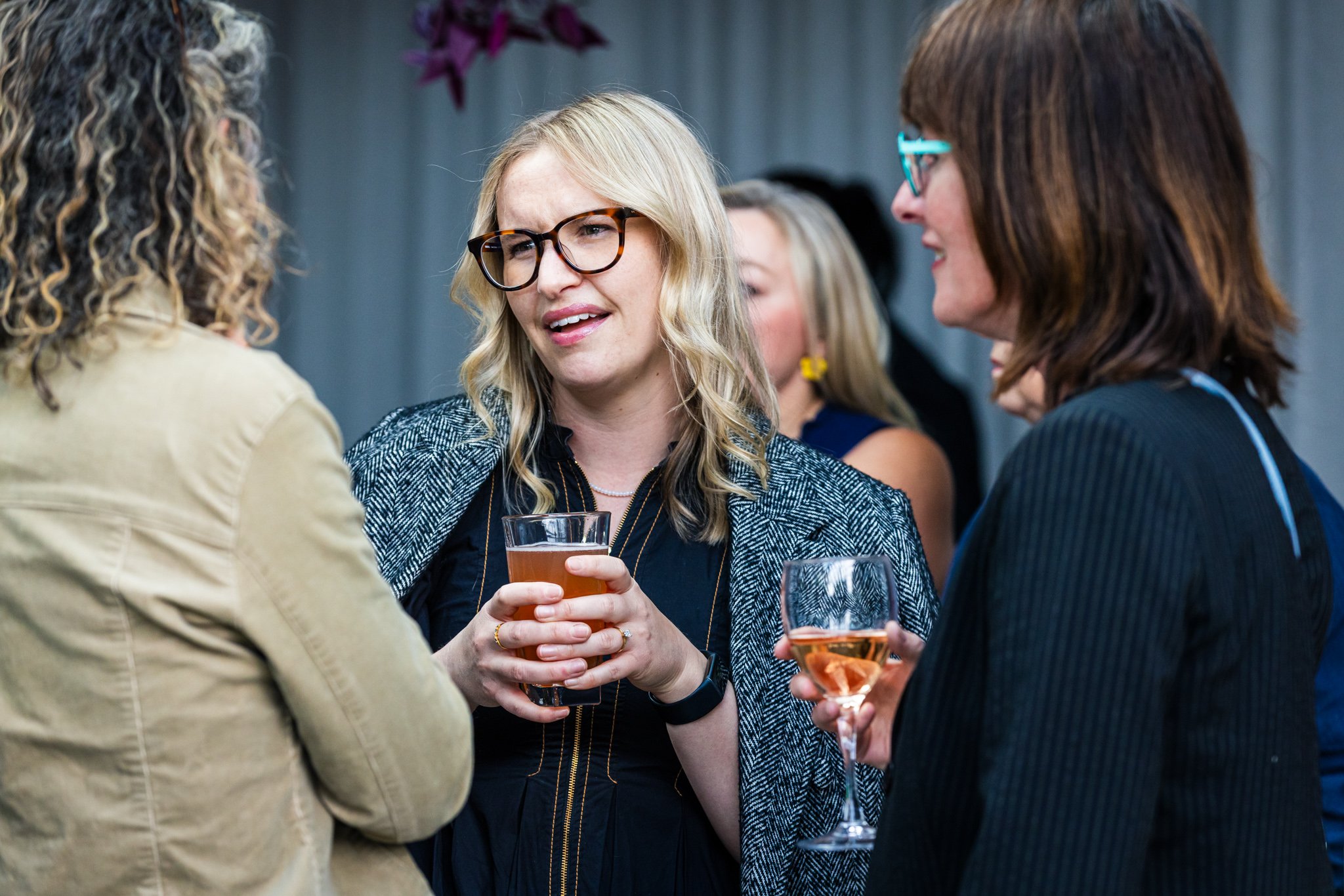 Three women engaged in conversation at a social gathering, holding drinks, with a blurred background.
