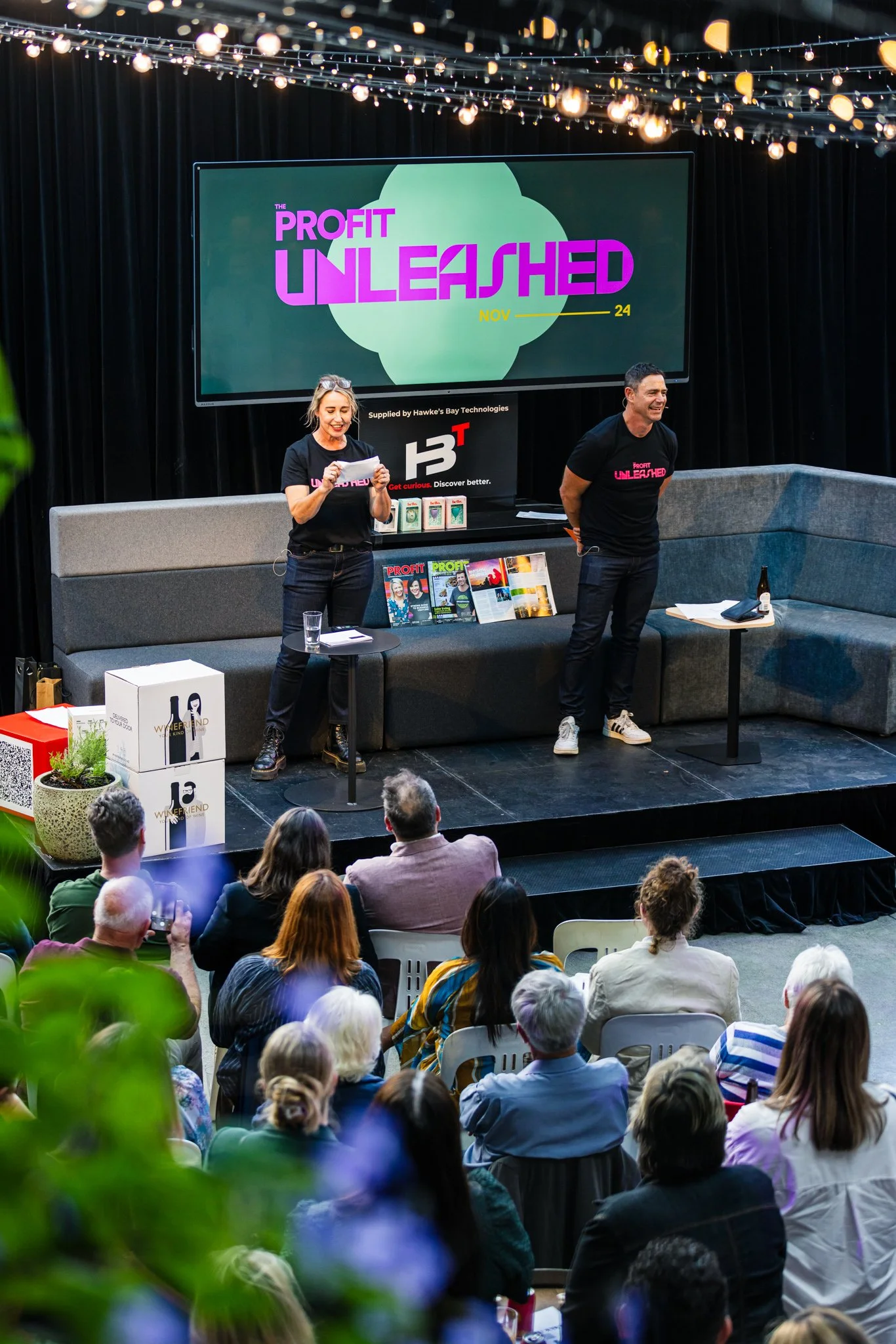 A presentation event with two speakers on a stage, one woman and one man, with a large screen behind them displaying 'Profit Unleashed' and the date November 24. The stage has a grey sofa, small tables, and magazines or catalogs. Audience members are