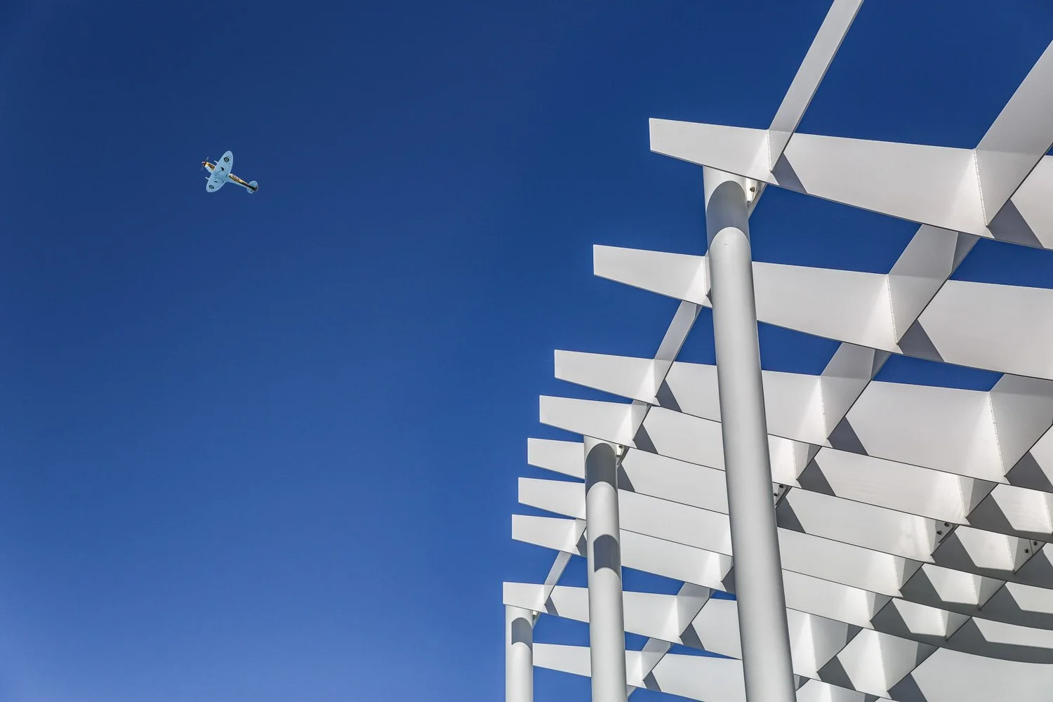 Modern building with white, sun-shaded panels and metallic poles against a deep blue sky, with a small propeller airplane flying in the background.